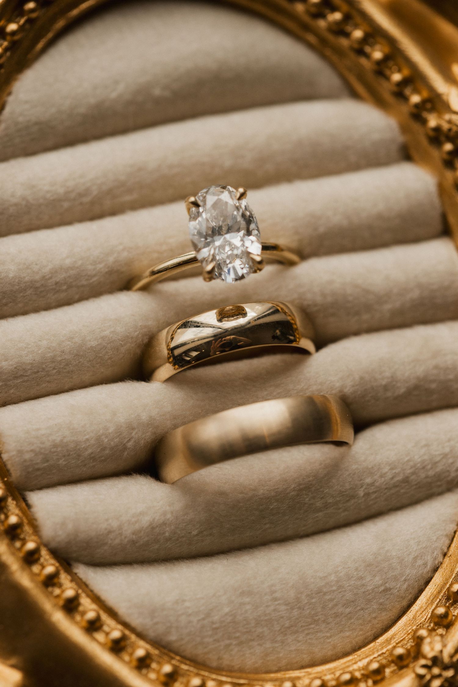 Gold oval diamond engagement ring and two gold wedding bands resting in a cream velvet ring box with an ornate gold frame.