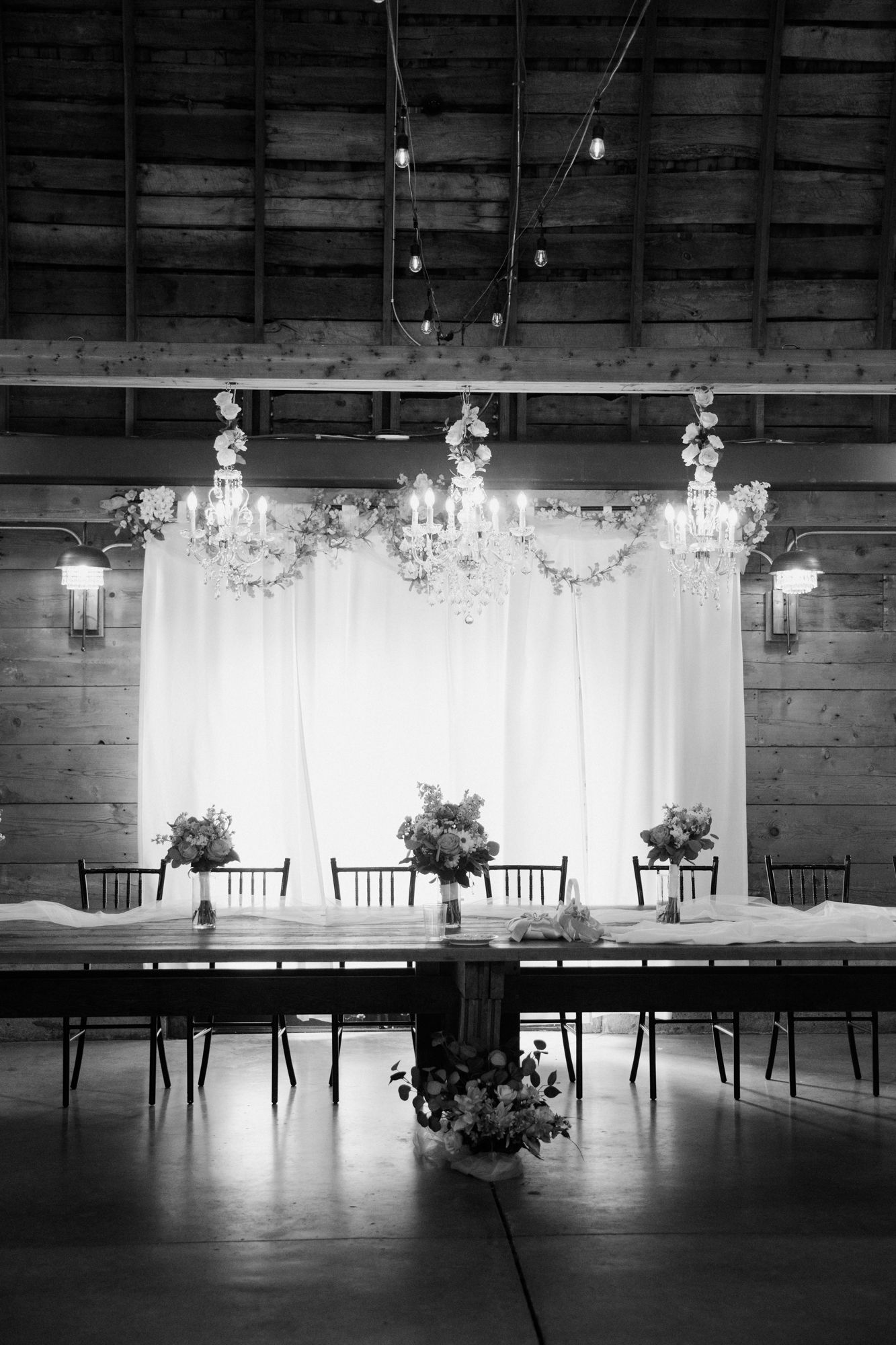 Black and white photo of elegant barn wedding reception with chandelier, floral arrangements, and draped white backdrop.