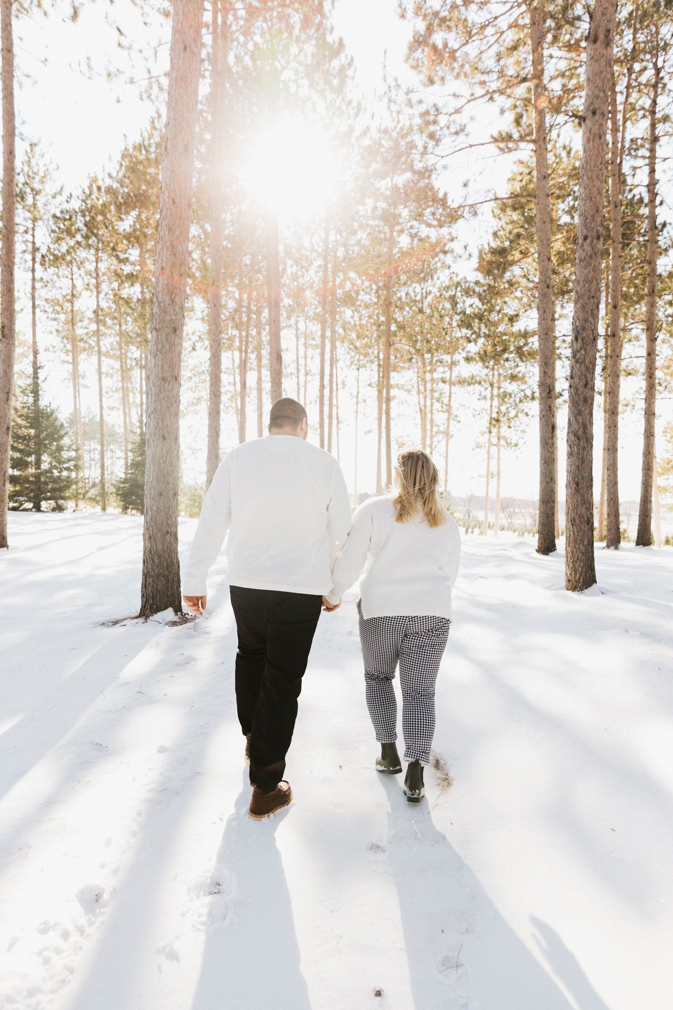 A couple walks hand in hand through a snowy forest backlit by winter sunlight.