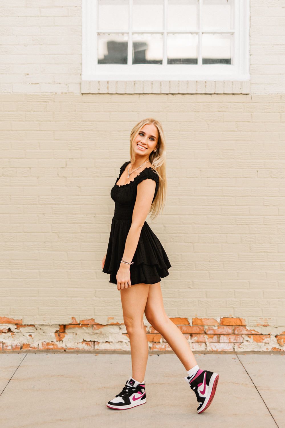 Playful pose wearing black dress and colorful sneakers against urban brick wall.