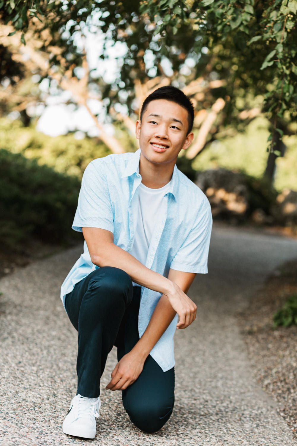 Casual pose on gravel path wearing blue shirt and dark pants with white sneakers.