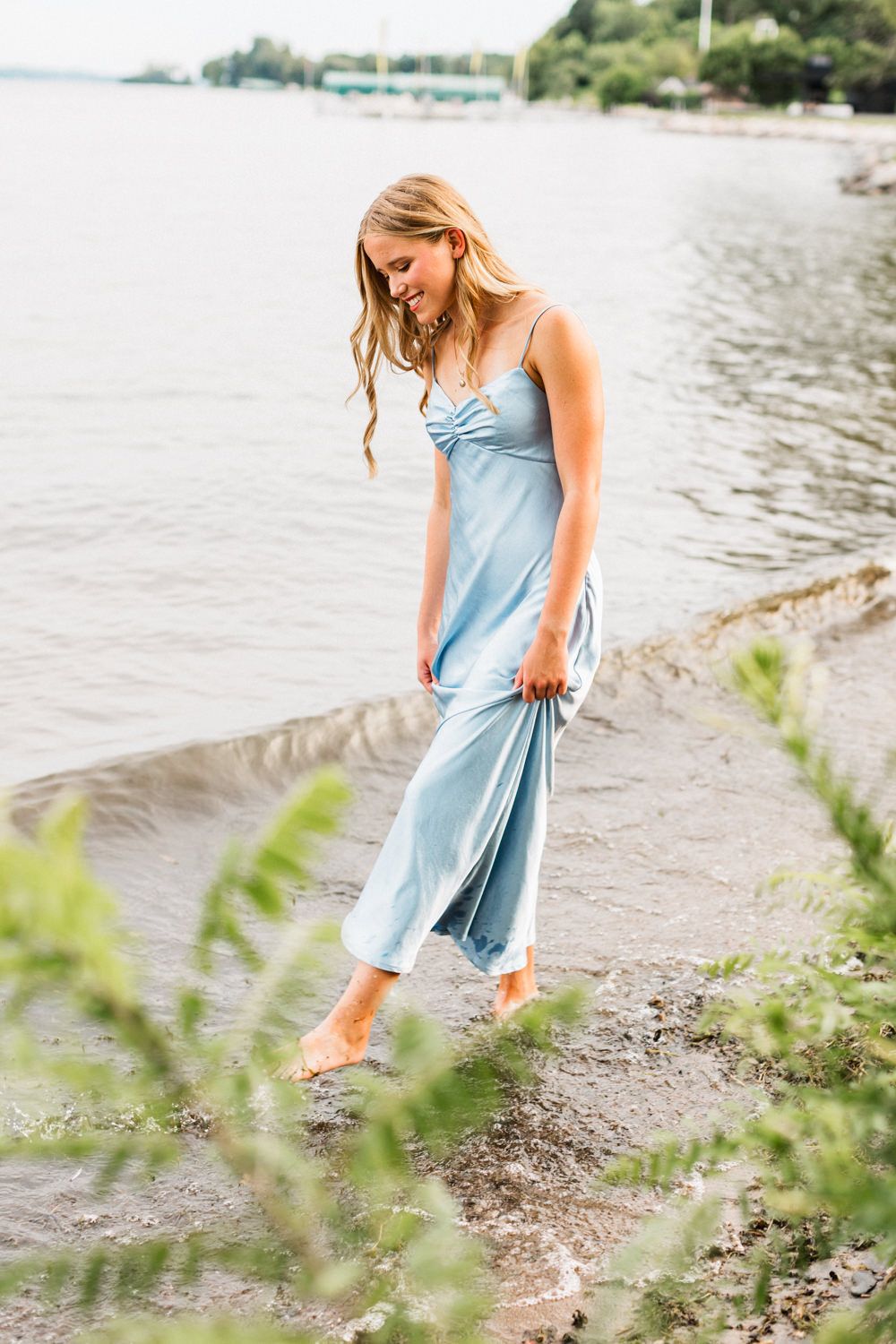 Woman in pale blue slip dress walks barefoot along waterfront path during summer photoshoot.