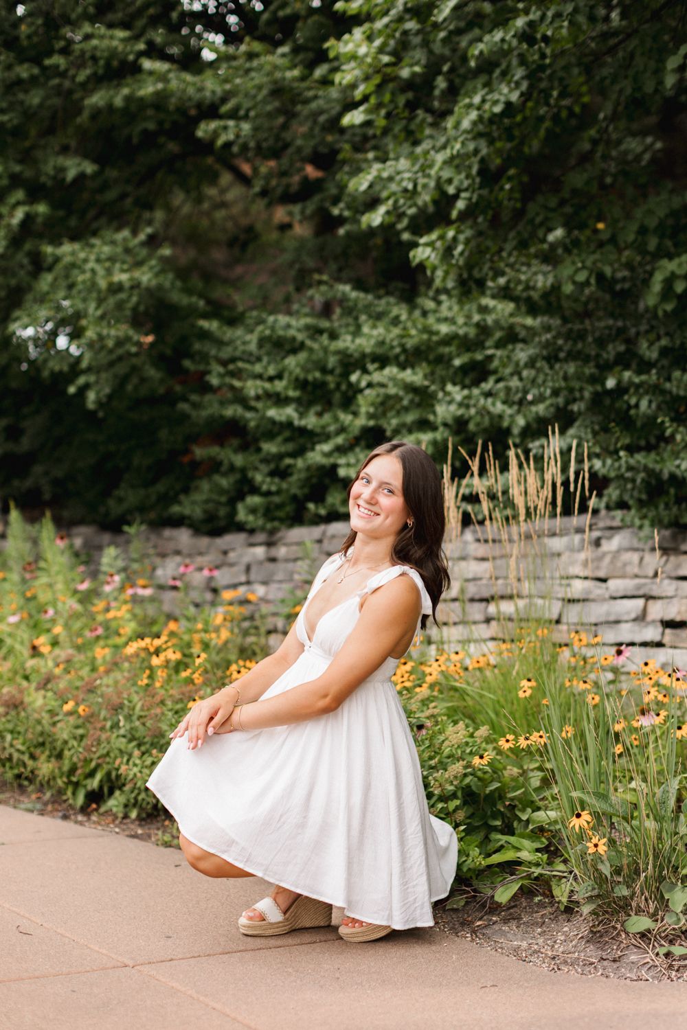 Figure in a flowing white summer dress poses gracefully against a garden stone wall with vibrant yellow flowers blooming.