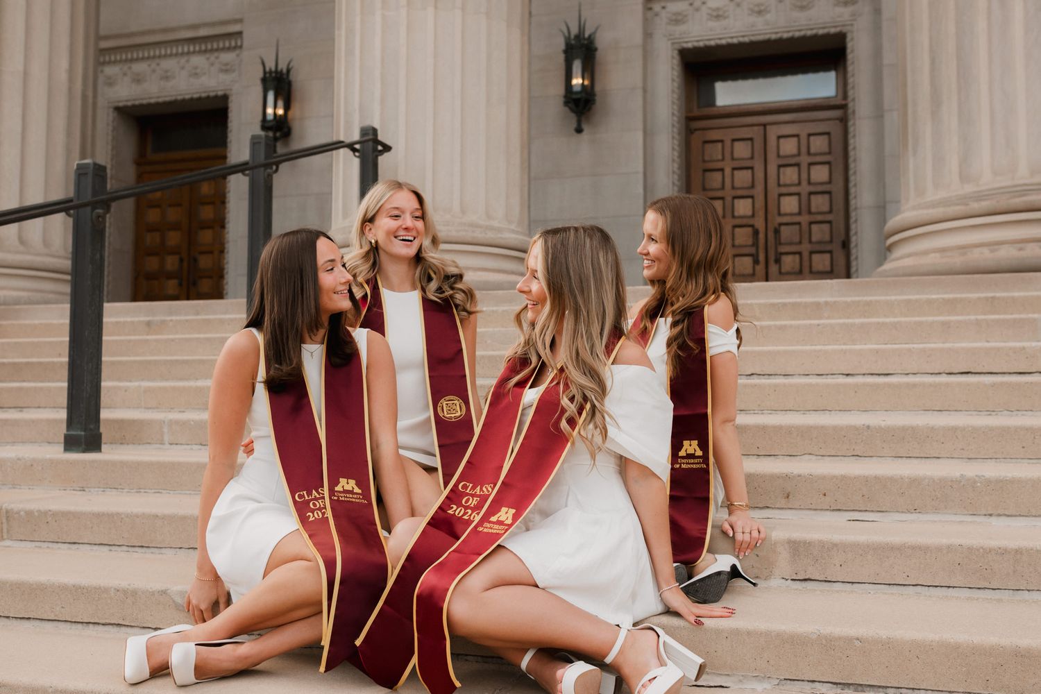 Four female University of Minnesota graduates in white dresses and maroon stoles sit laughing on steps.