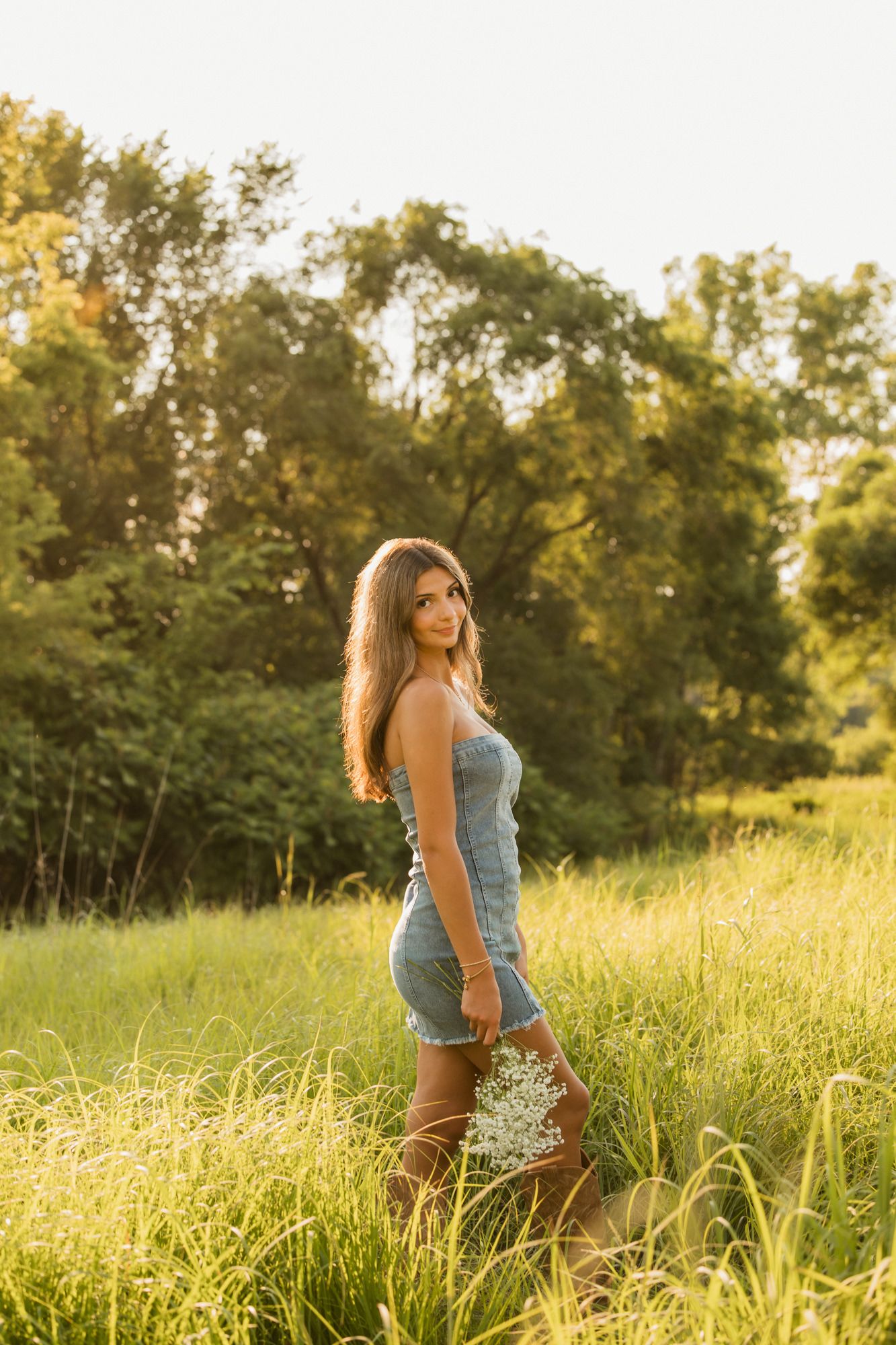 Young woman in denim dress holding wildflowers stands in sunlit green meadow with trees in golden hour light.
