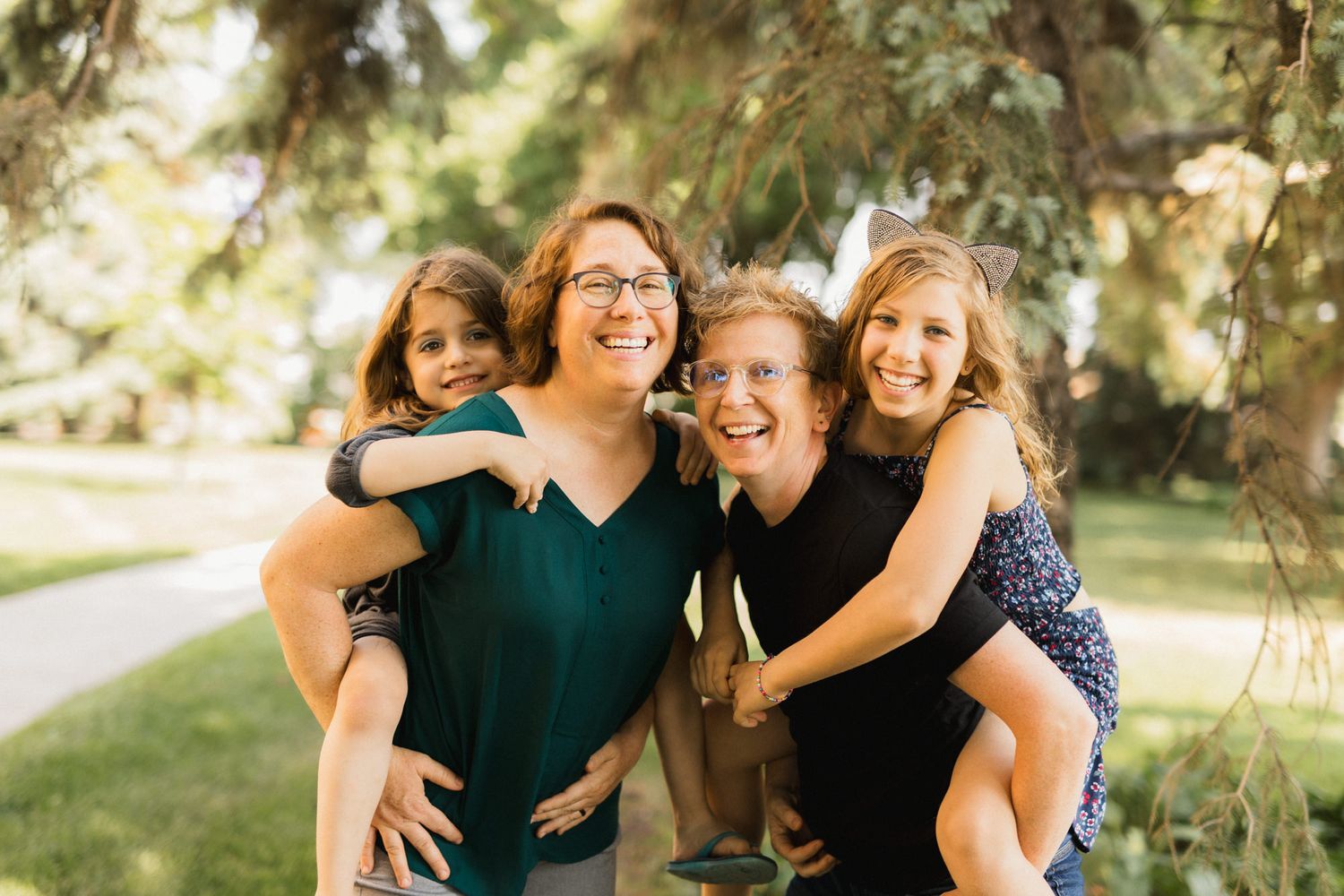 Two women giving piggyback rides to two girls, smiling outdoors under green trees in a warm family portrait.