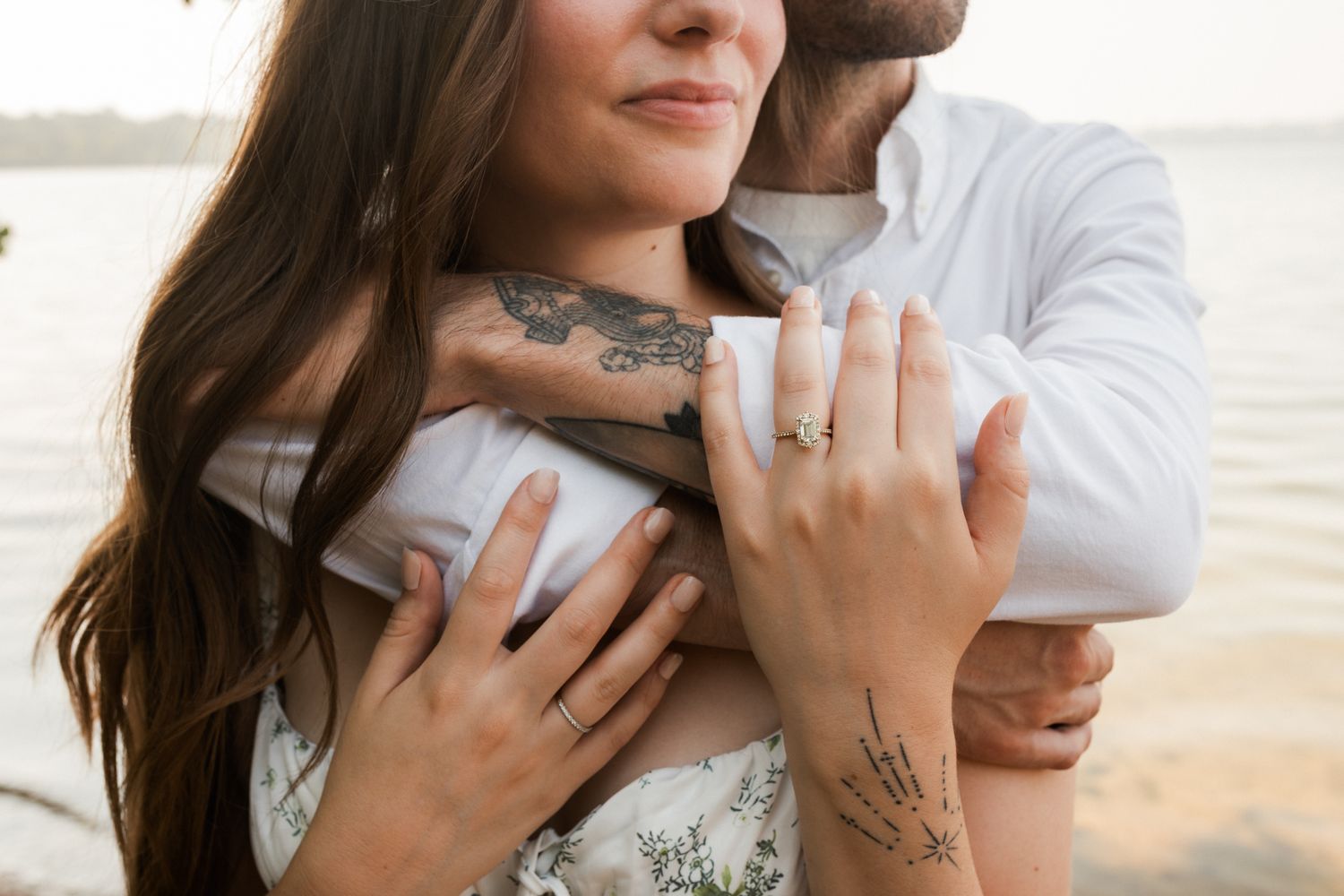Couple embracing near water, woman showing engagement ring, both with tattoos, romantic outdoor engagement photo.