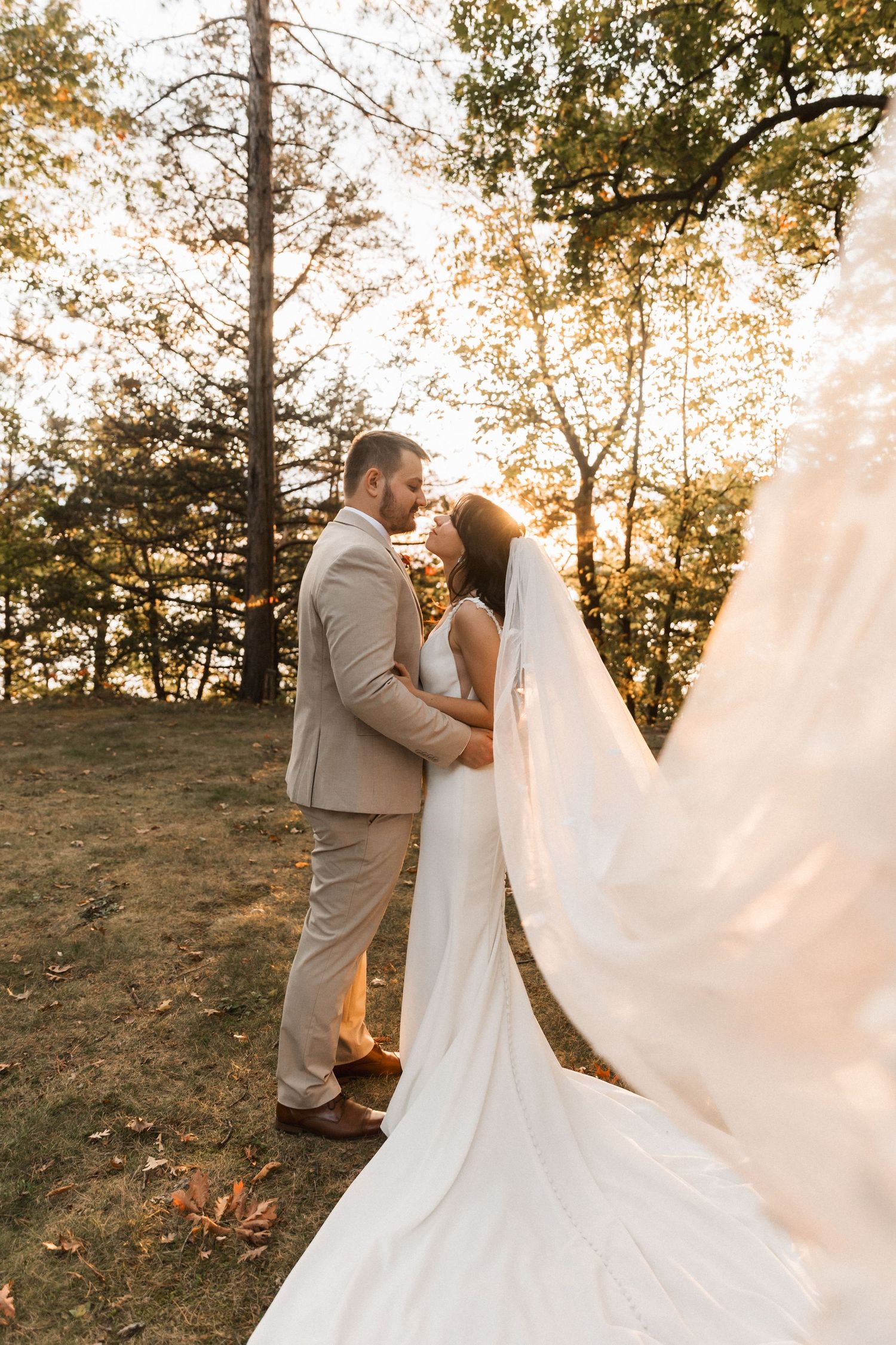 Romantic wedding couple embraces during golden hour sunset in a wooded outdoor setting with flowing veil.