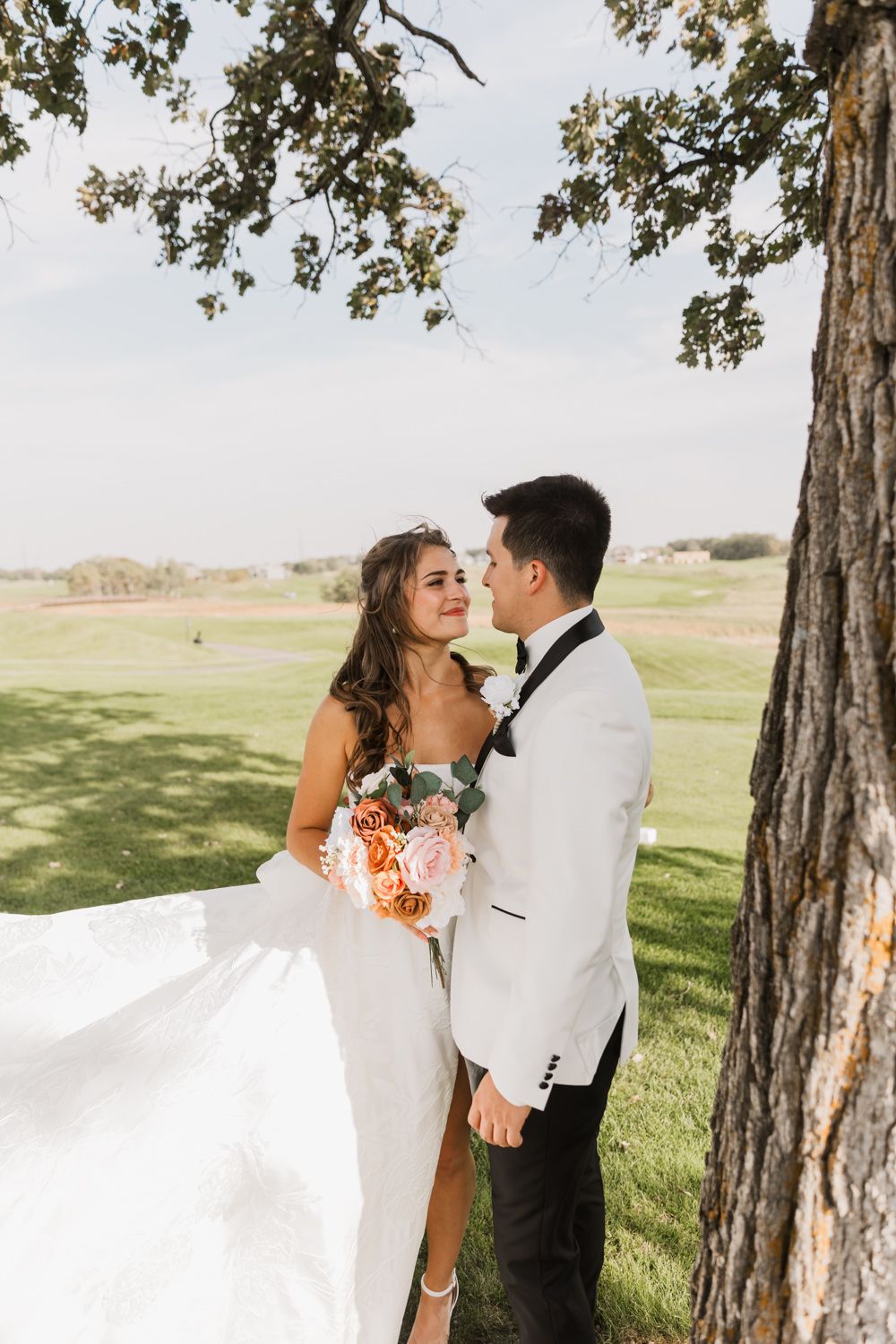 A romantic wedding photo captured under a tree on a golf course with a flowing white dress and coral bouquet.