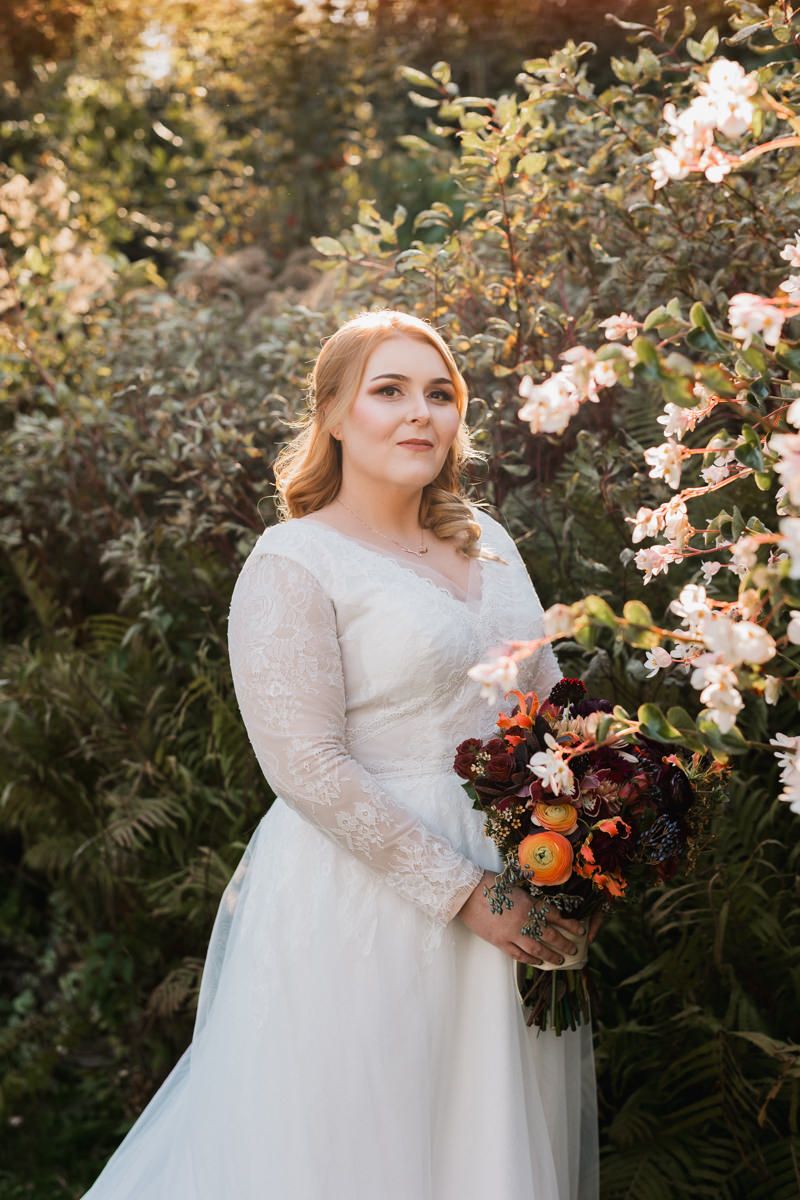 A bride in a lace-sleeved white wedding dress poses outdoors among flowering bushes with a colorful autumn bouquet.