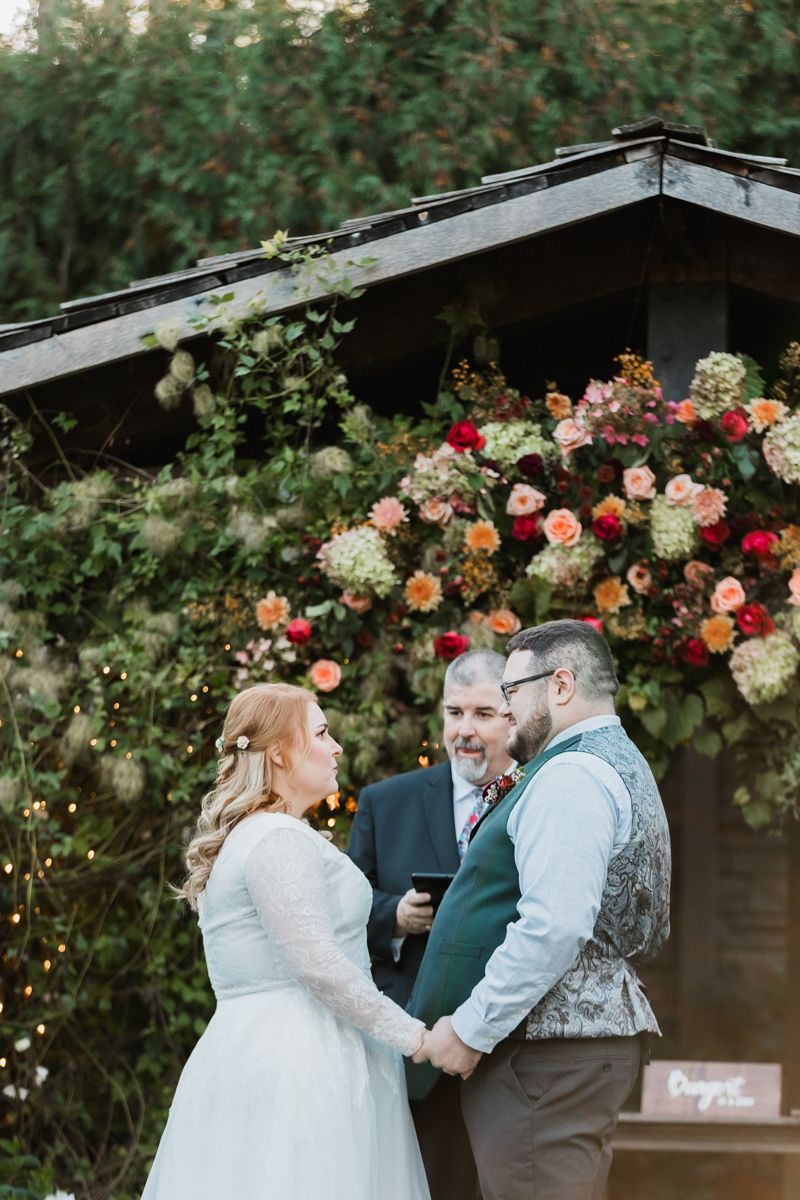 Wedding ceremony takes place under rustic wooden pergola adorned with colorful floral arrangements.