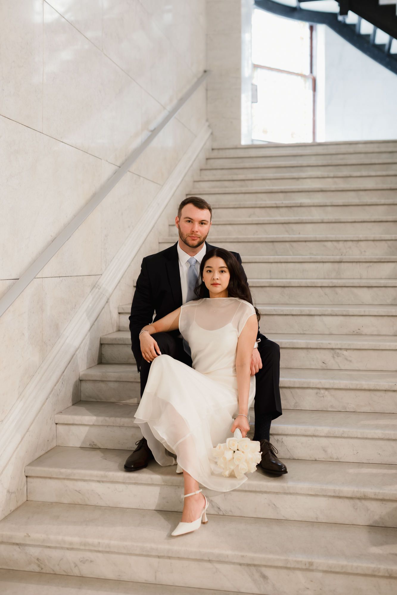Bride and groom sitting on marble stairs, she in white dress with bouquet, he in black suit, elegant wedding portrait.