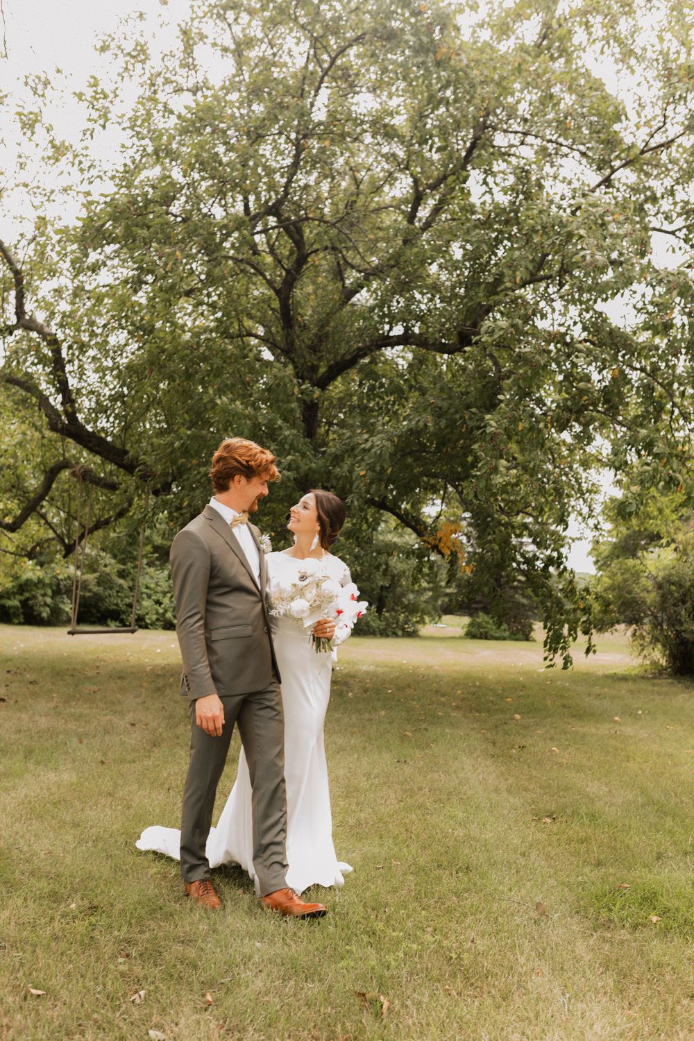 A couple in wedding attire dances playfully in a sunlit meadow beneath a large tree.