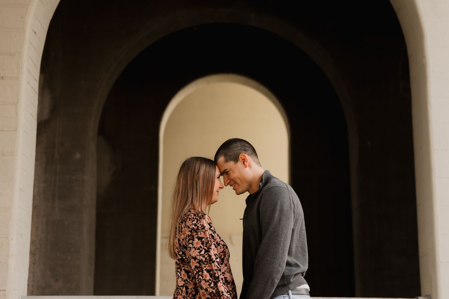Romantic couple touching foreheads under elegant arched doorways in soft warm lighting.