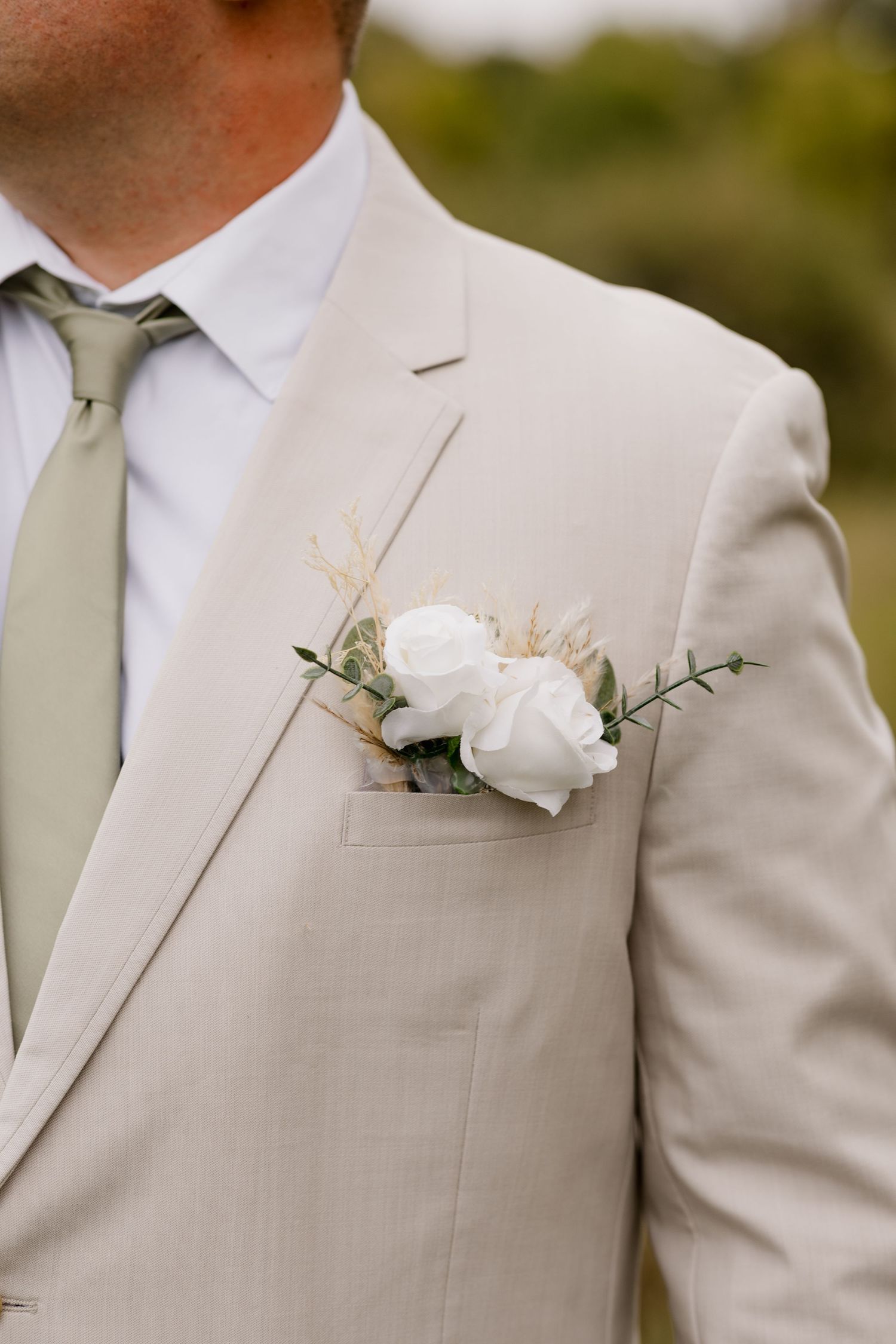 Groom in beige suit with sage green tie and white rose boutonniere with eucalyptus and pampas grass.
