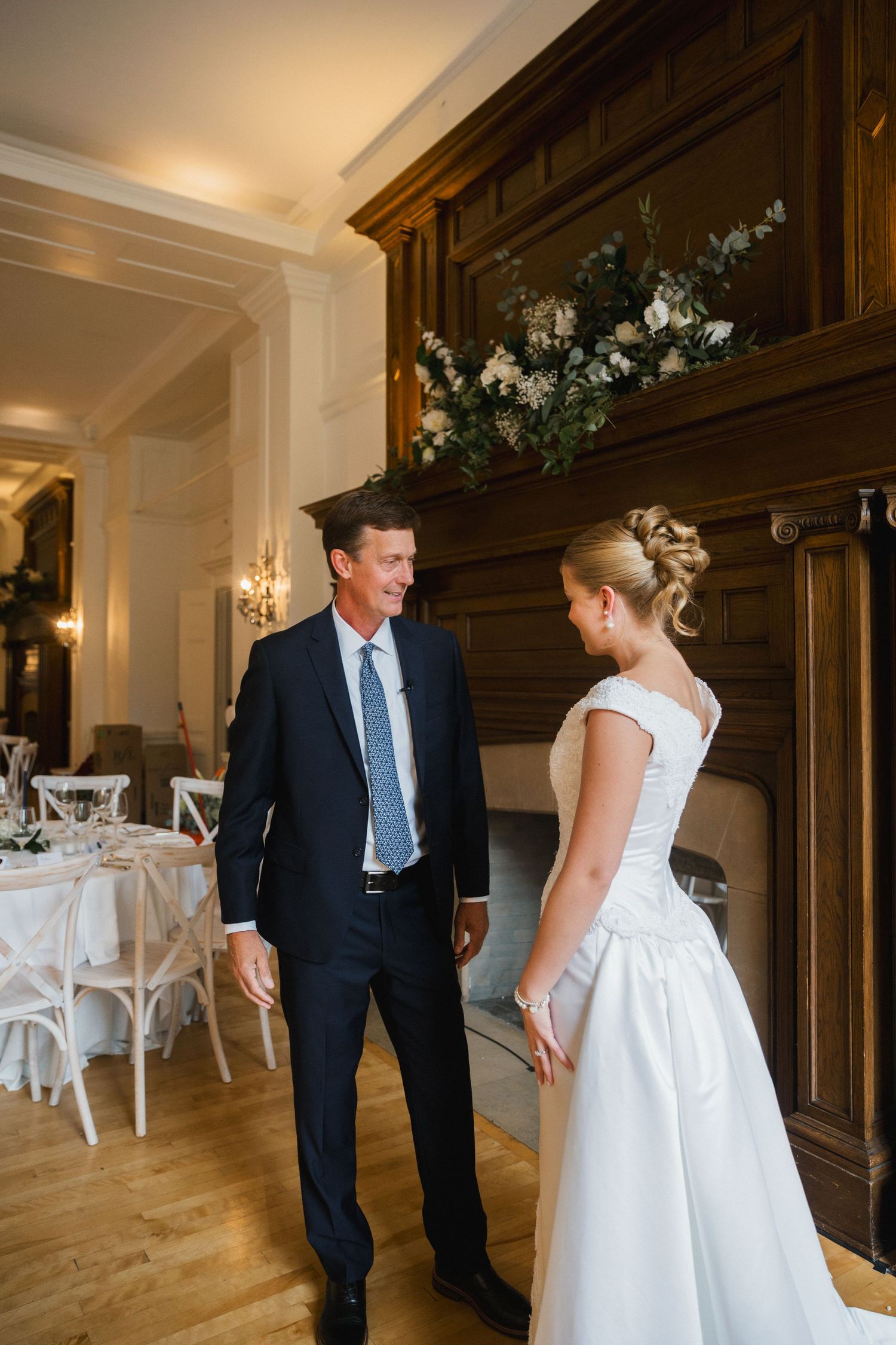Bride in white gown shares a joyful moment with a man in a navy suit near an elegant fireplace in a wedding venue.