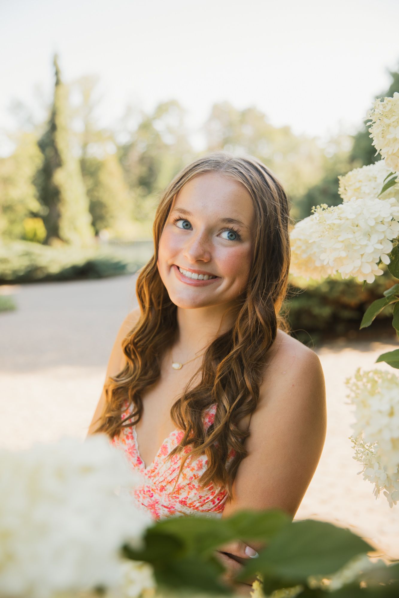 Young woman with long wavy hair smiling among white hydrangea blooms in a sunny garden wearing a floral dress.