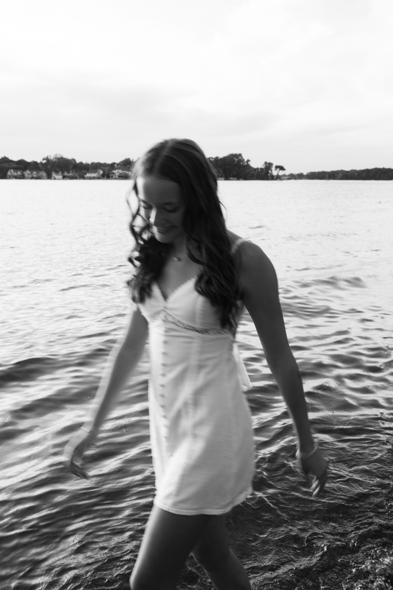 Black and white photo of a young woman in a white dress wading in calm lake water near a shoreline.