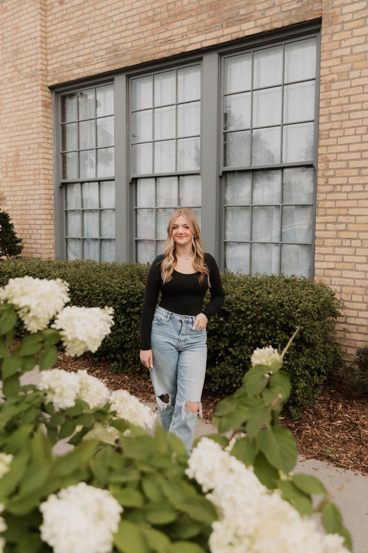 Smiling blonde woman in black top and jeans posing near white hydrangeas outside brick building.