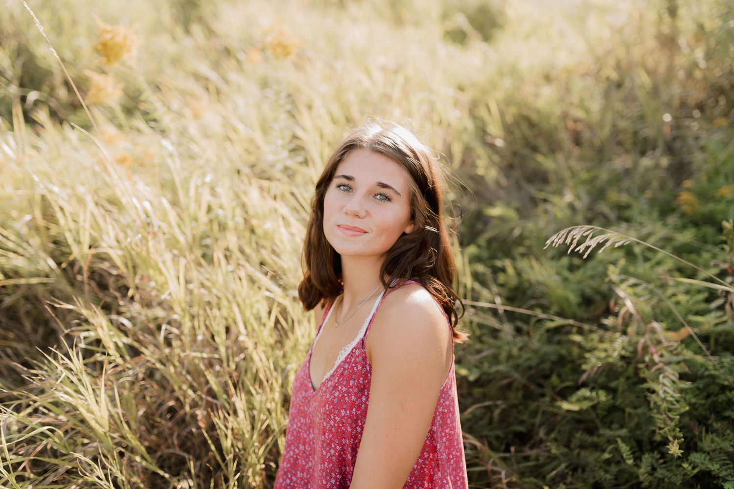Young brunette woman in pink floral dress posing in golden sunlit tall grass field for portrait photo.
