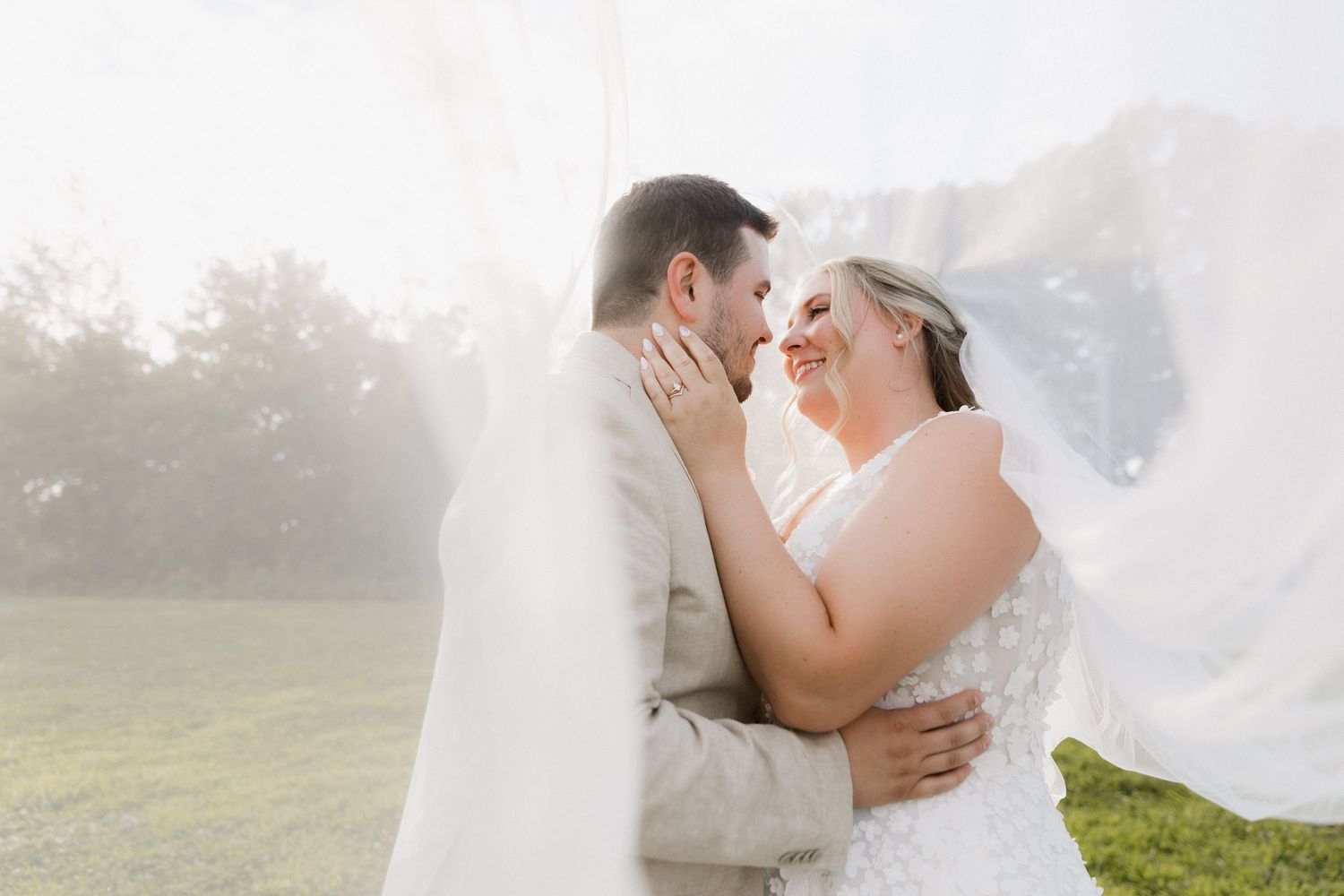 Bride and groom share a romantic moment under a flowing white veil outdoors on their wedding day.