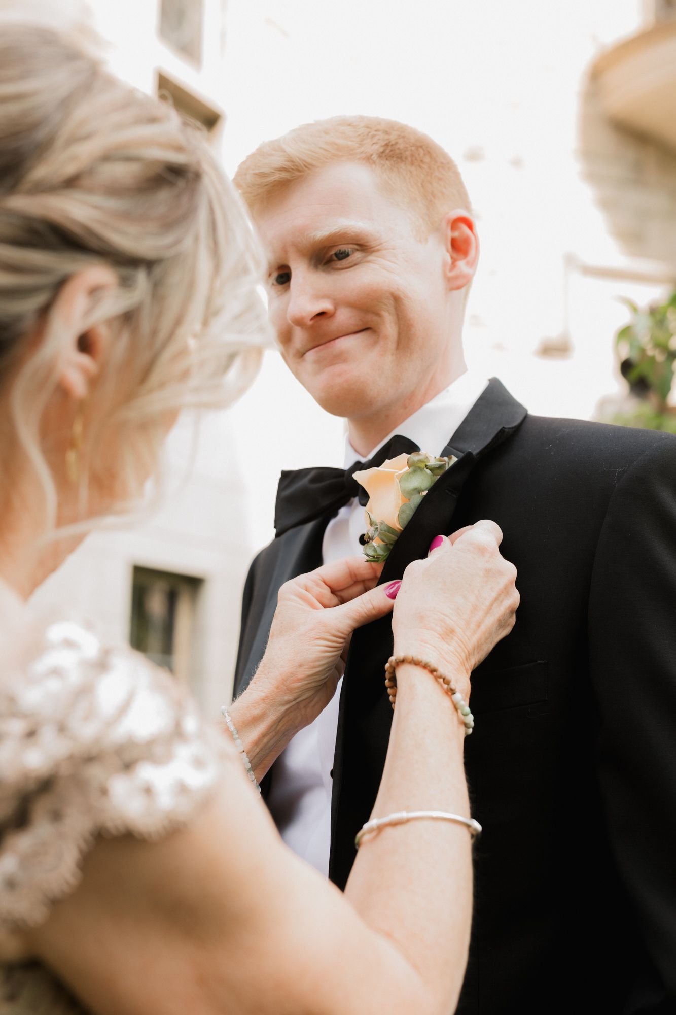 A smiling groom in a black tuxedo has a floral boutonniere pinned to his lapel by a woman in lace.