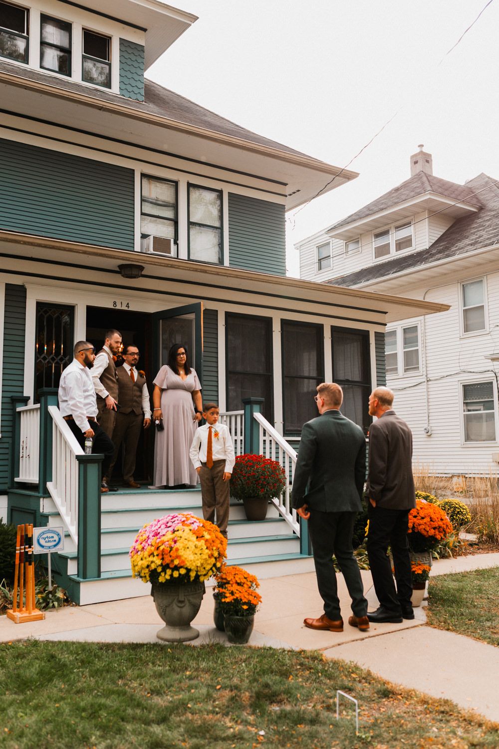 People gather outside a green house decorated with orange and yellow fall flowers and pumpkins for a seasonal celebration.