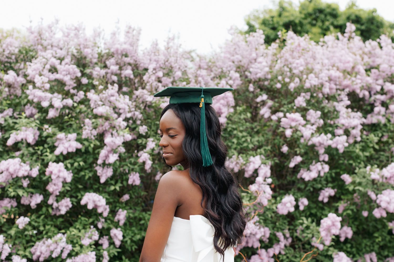 Graduate in strapless dress poses in front of pink flowering bushes.