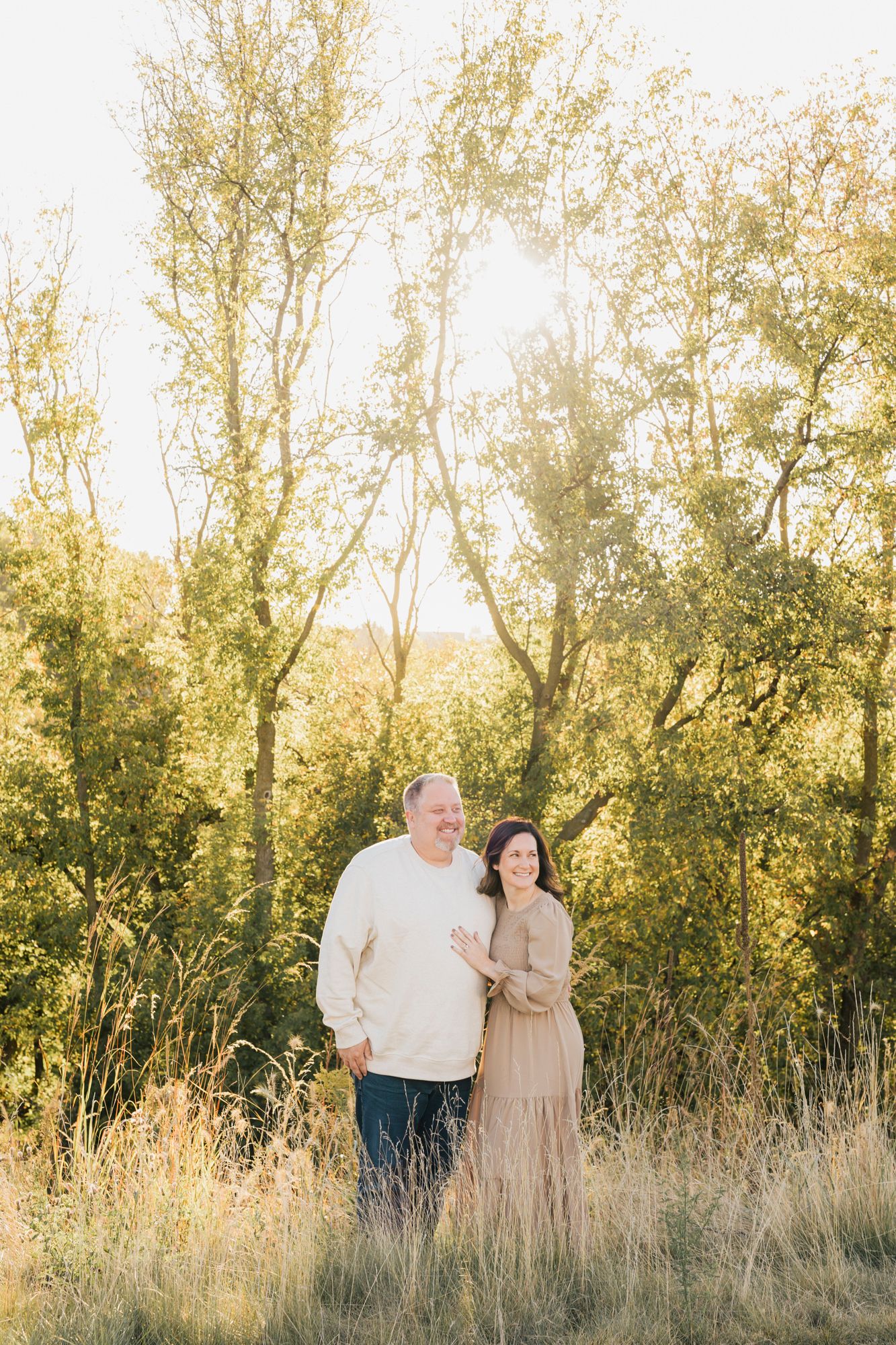 A romantic portrait of a couple standing in golden sunlit grass with autumn trees behind them.