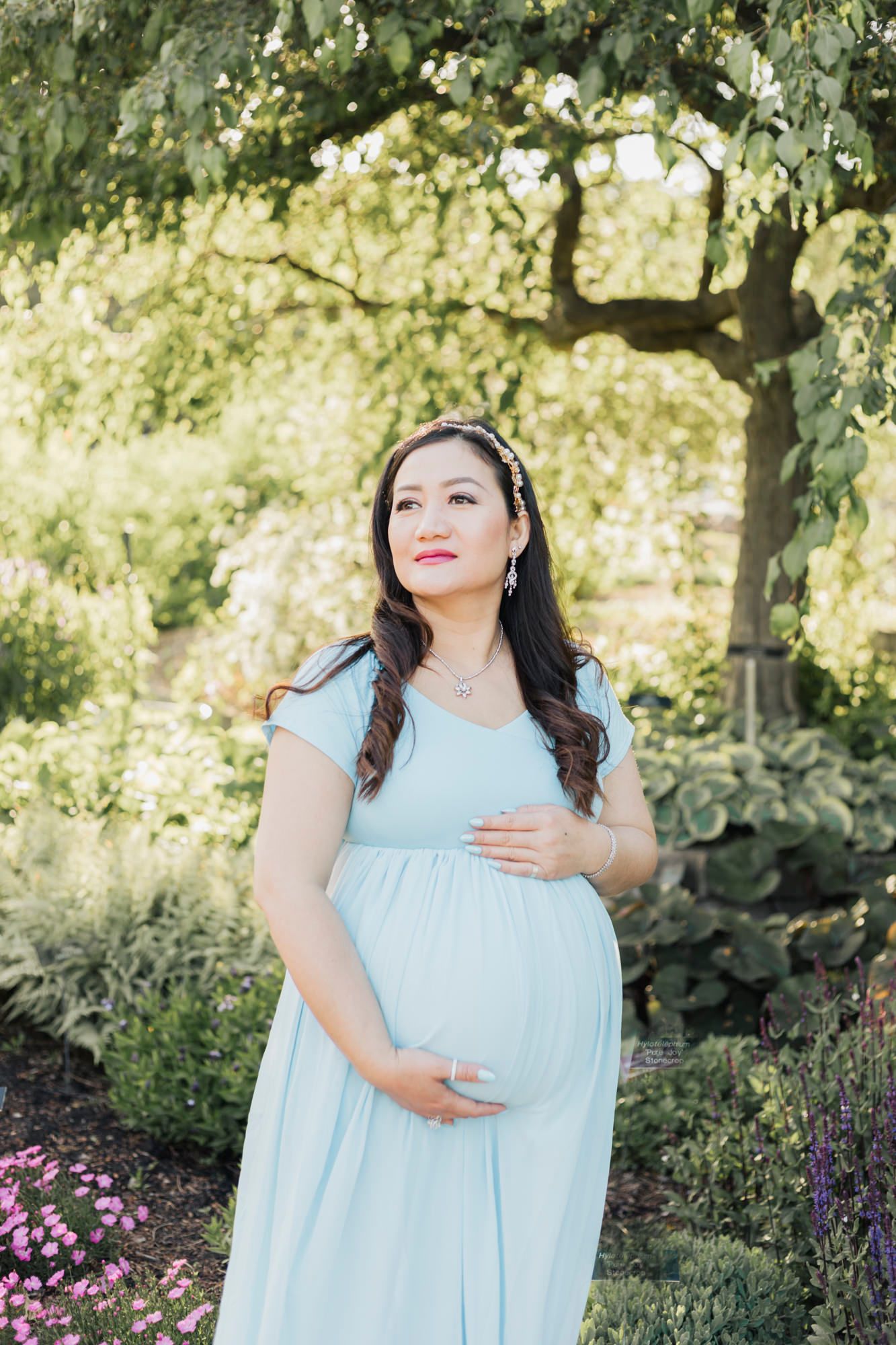 Woman in light blue dress poses gracefully in a garden setting with blooming flowers.