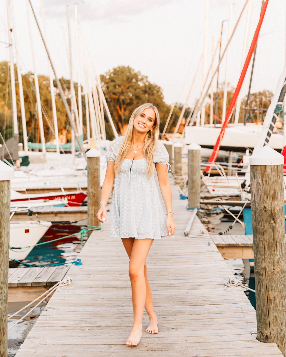 Woman in light summer dress walking barefoot along marina boardwalk at sunset.