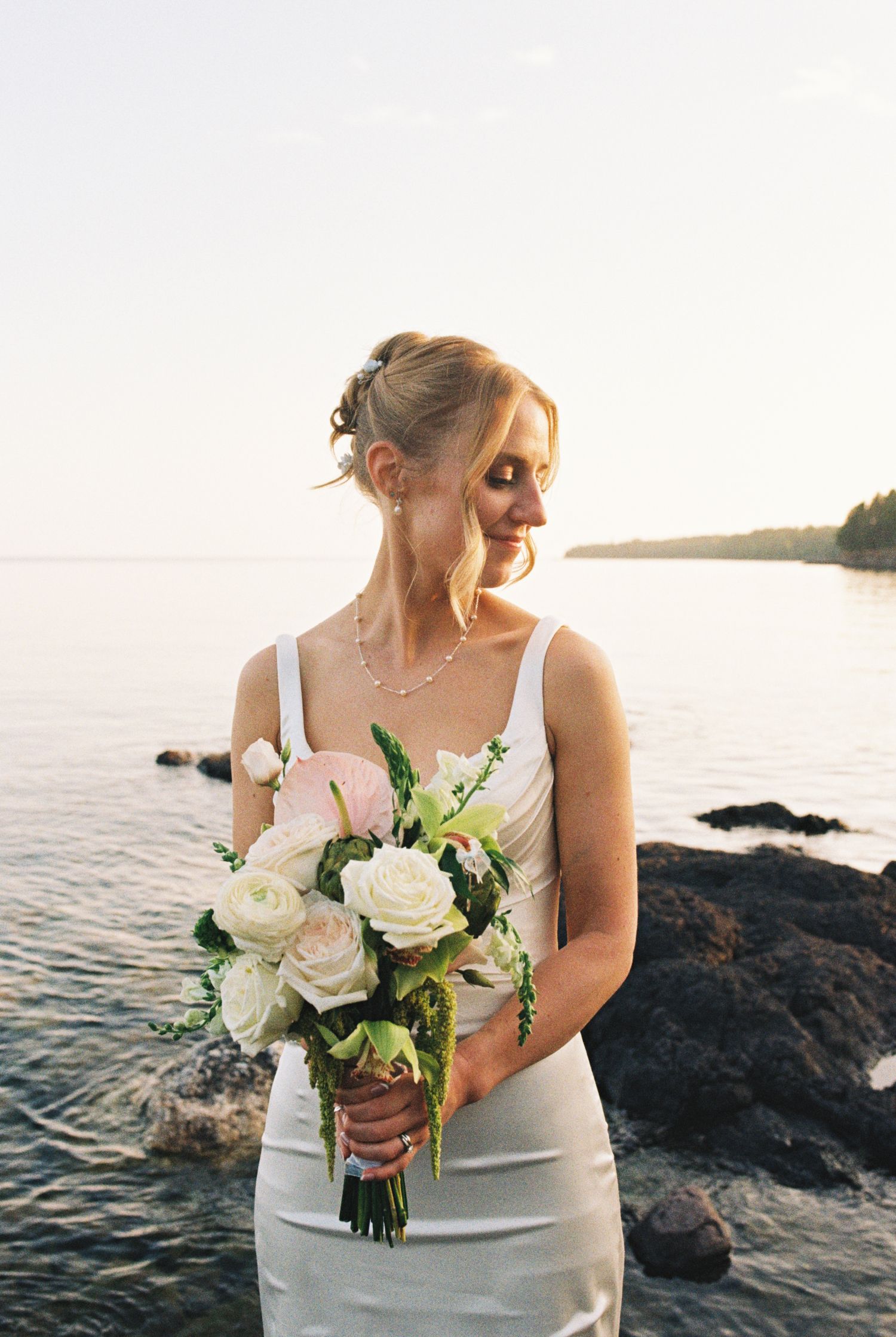 Bride in white gown holds pink and white bouquet by rocky shoreline at sunset.