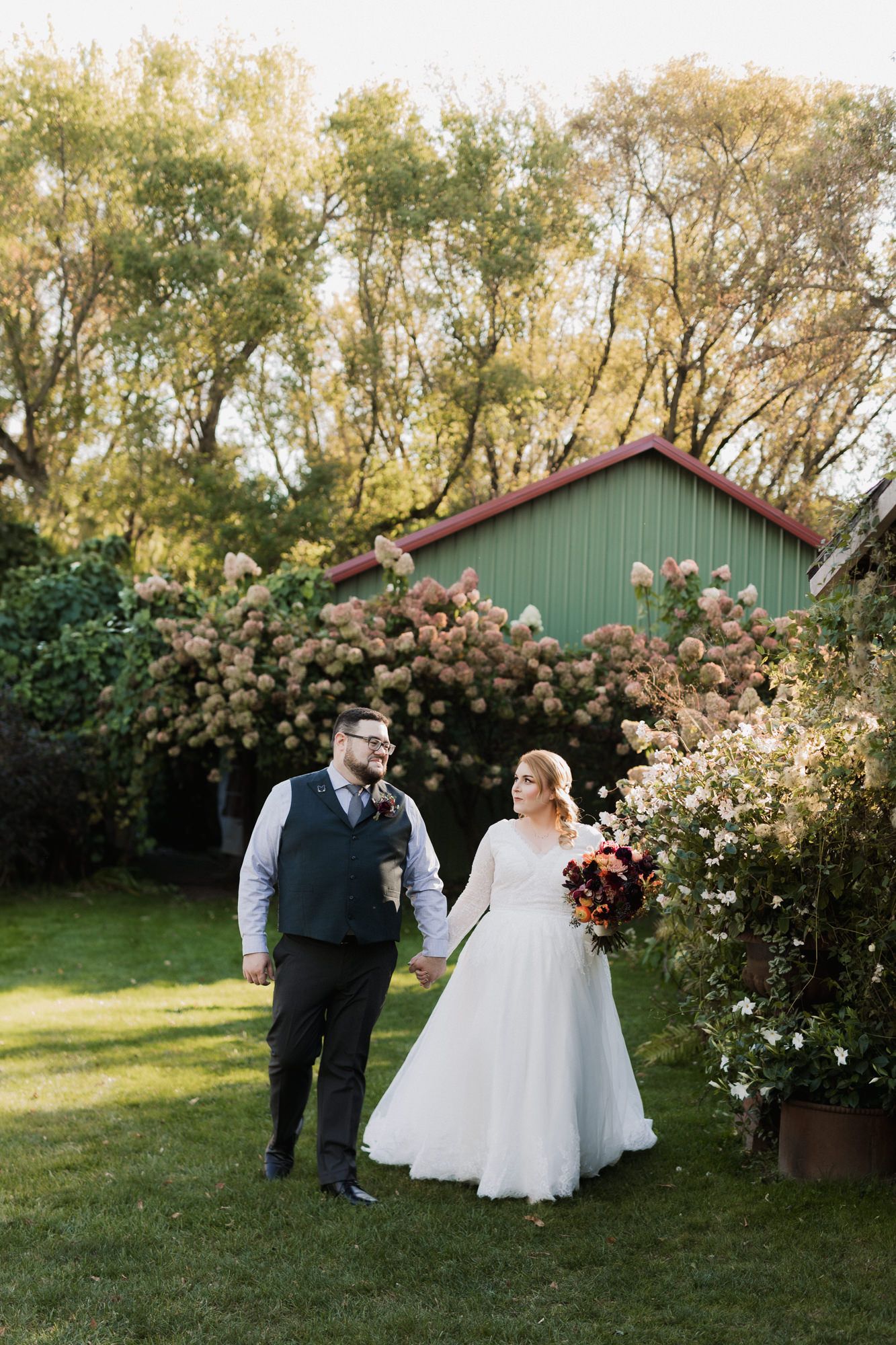 Bride and groom holding hands in lush garden with blooming flowers and green barn backdrop on wedding day.