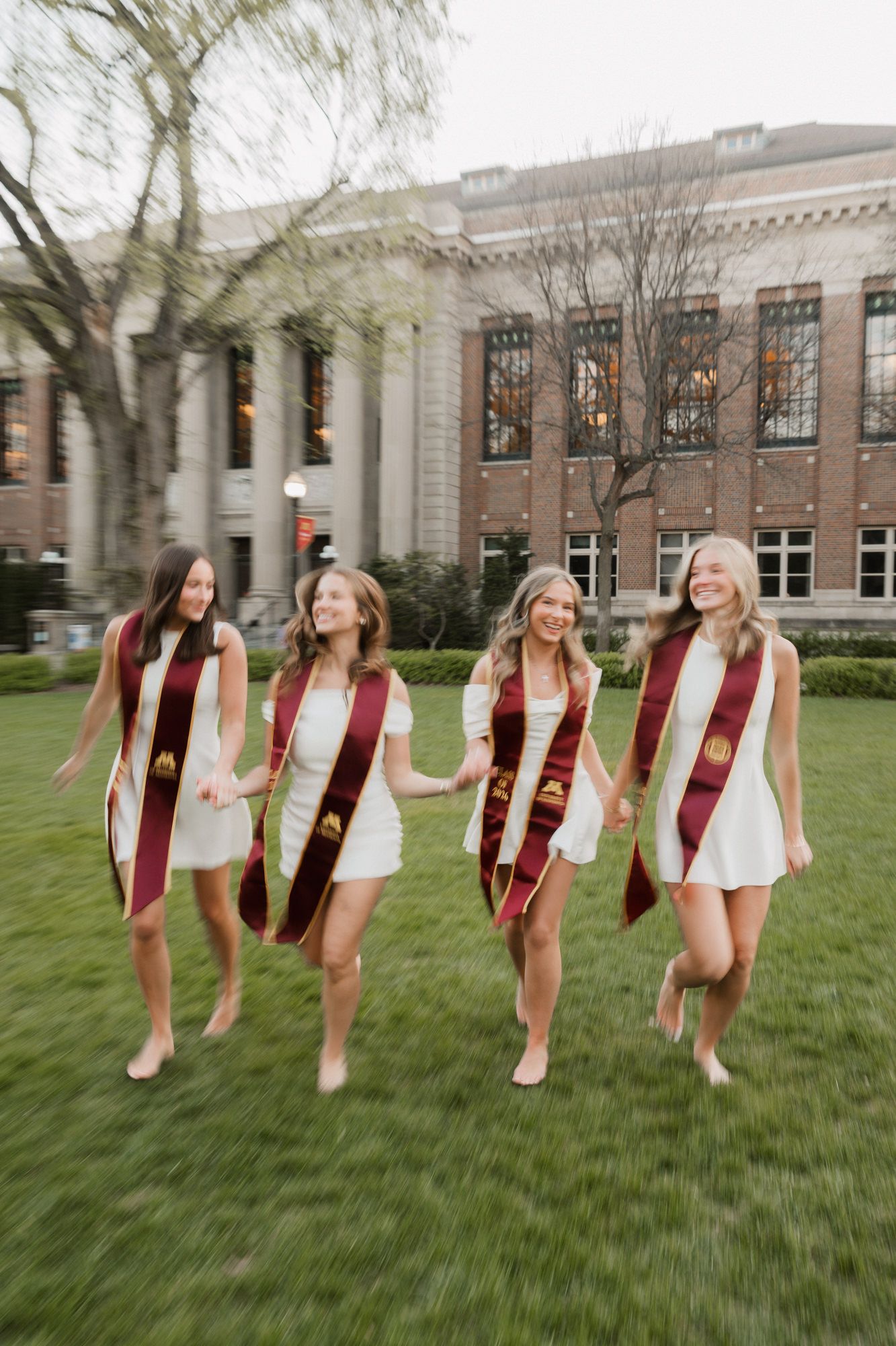 Four college graduates in white dresses and maroon graduation sashes walk barefoot across green campus lawn.