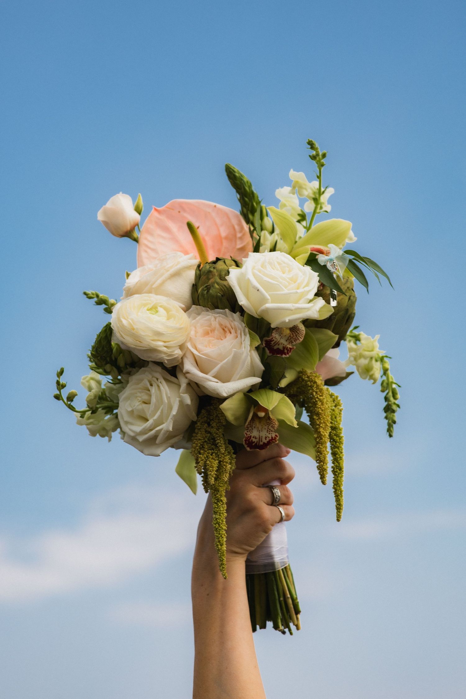 Hand holding a bridal bouquet of white roses, pink flowers, and greenery against a blue sky.