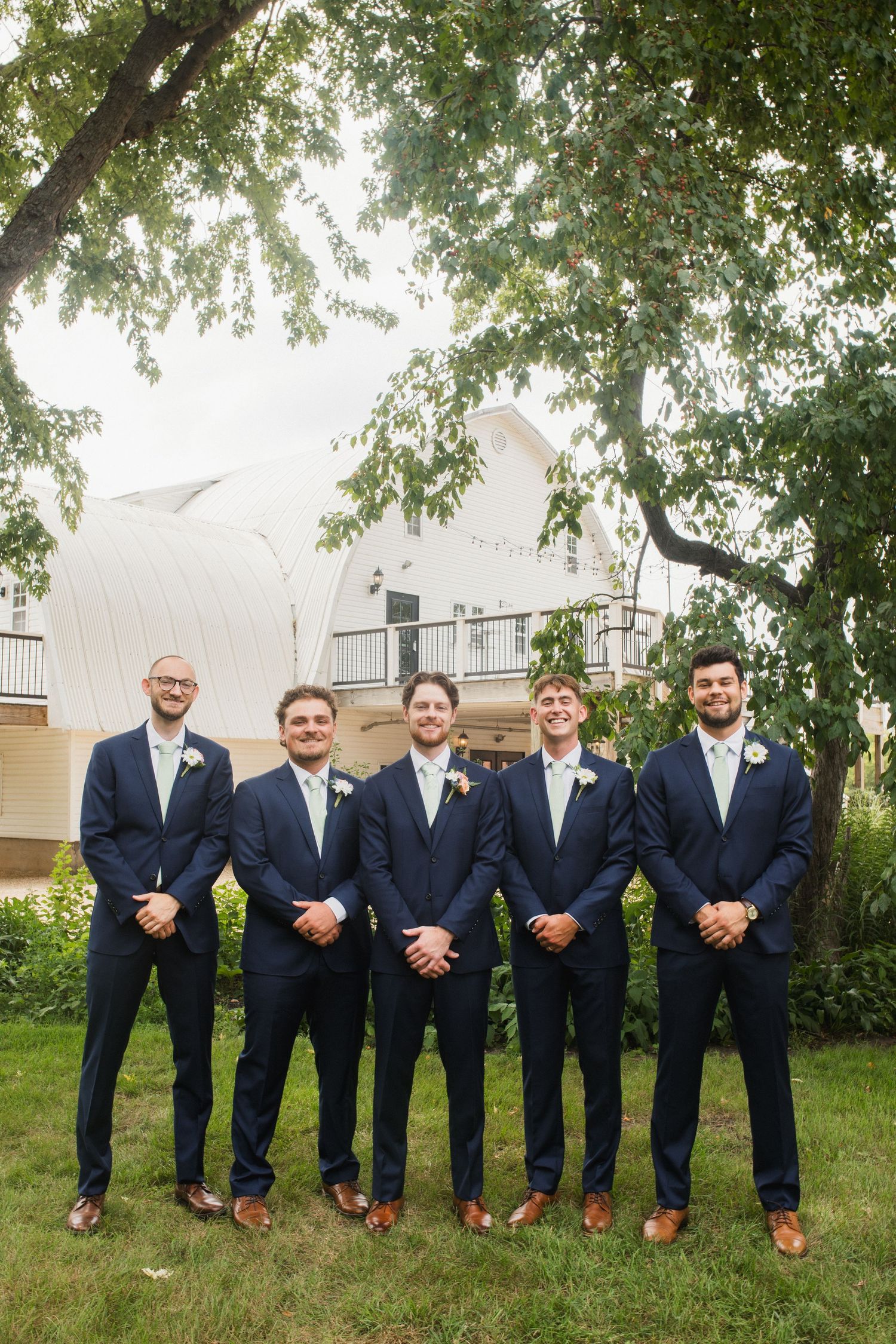 Five groomsmen in navy suits with light green ties and boutonnieres posing outdoors on grass near a white building.