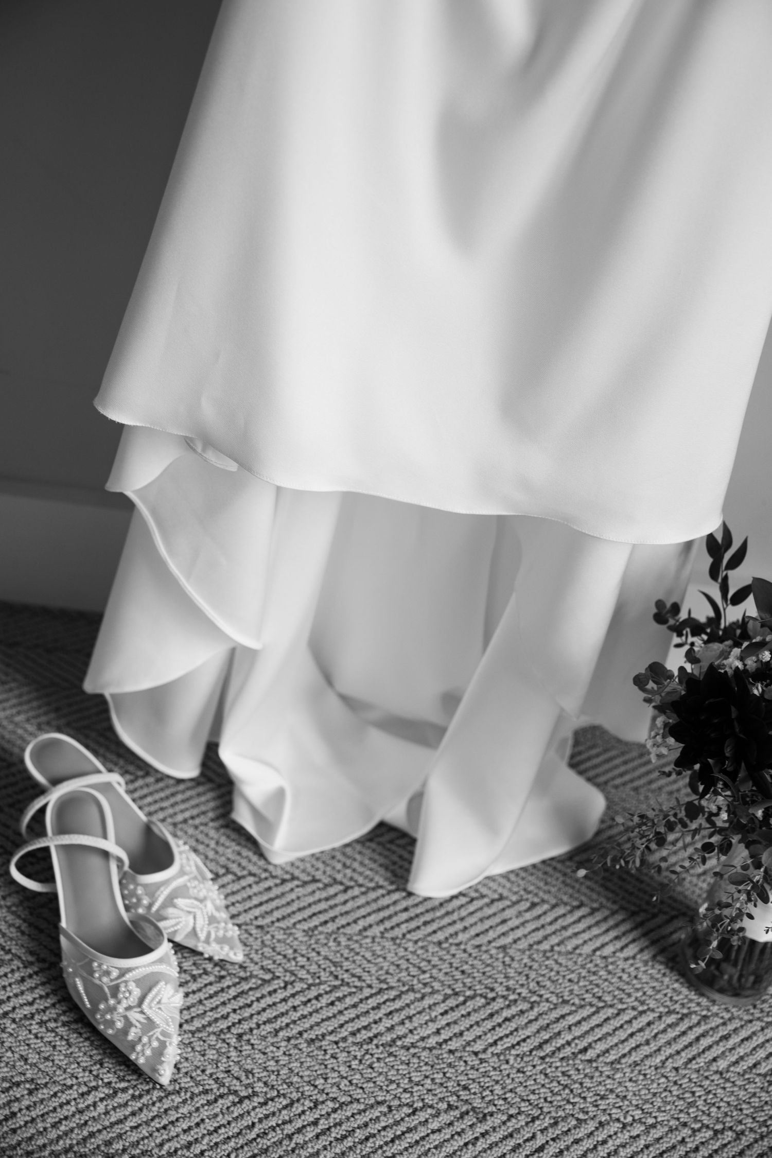 Black and white photo of a wedding gown hanging above beaded bridal heels and a small bouquet on a textured carpet.