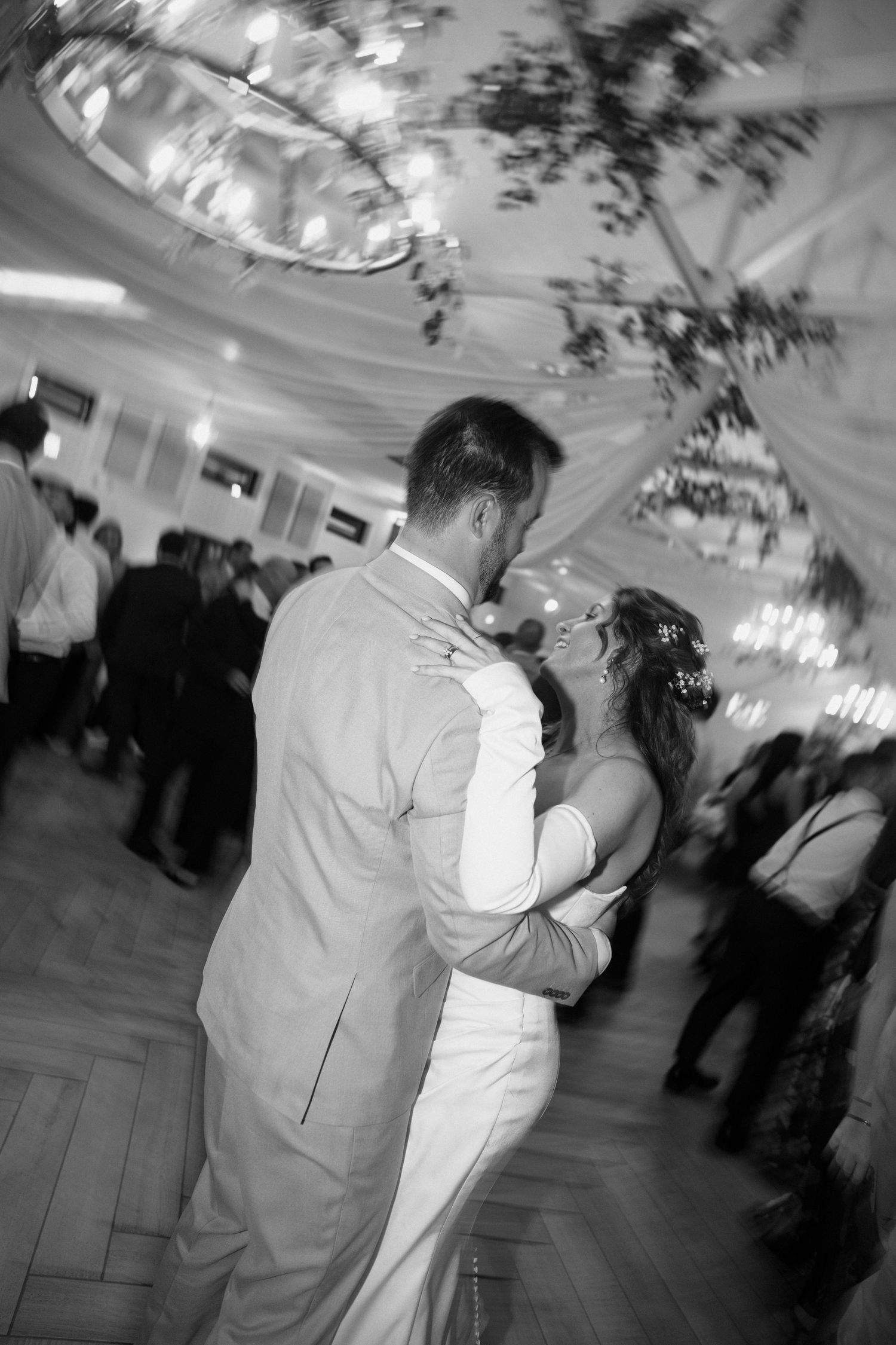Black and white photo of a bride and groom sharing a close dance on a wooden floor beneath chandeliers as guests look on.
