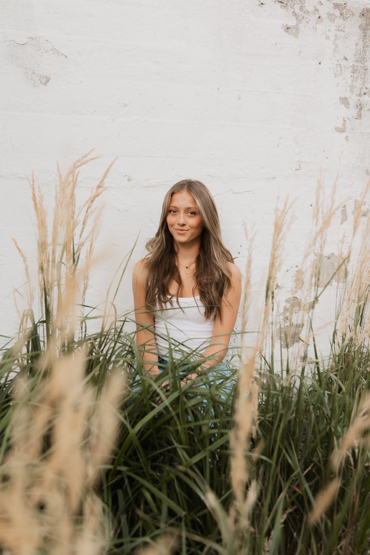 Young woman in white tank top sitting among tall ornamental grasses against white brick wall outdoors.