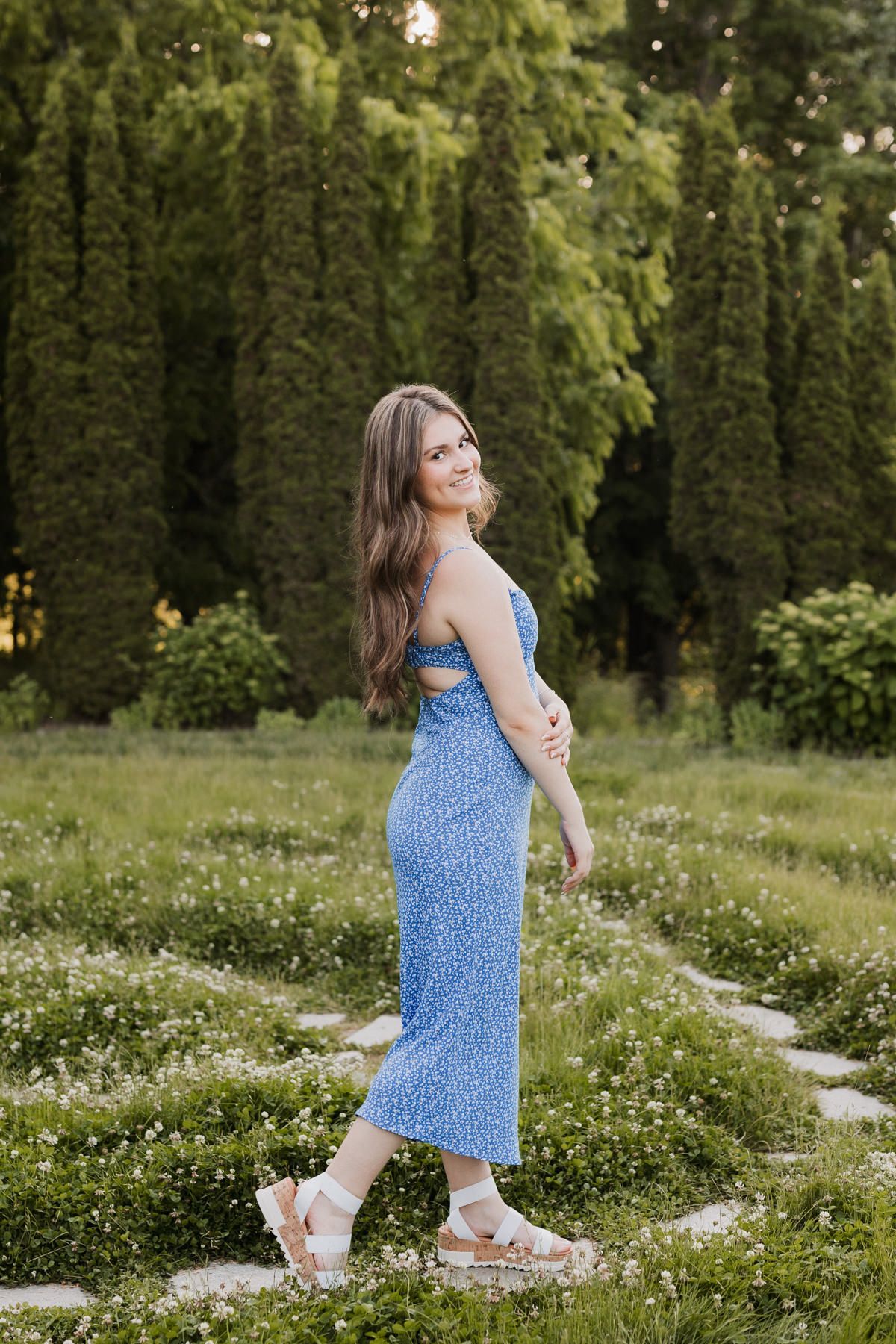 Young woman in blue floral midi dress walking on garden path with tall evergreen trees in background.