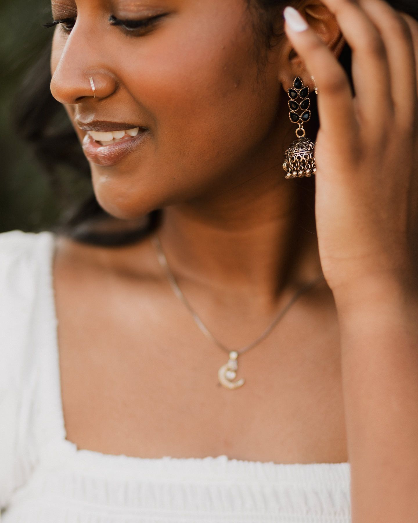 Young woman smiling wearing ornate gold jhumka earrings and a delicate pearl pendant necklace in white top.