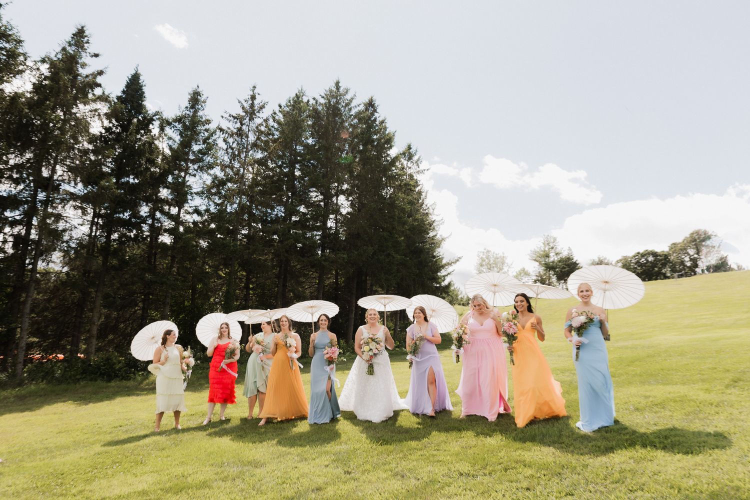 Bride and bridesmaids in colorful dresses holding white parasols on a green lawn with tall trees behind them.