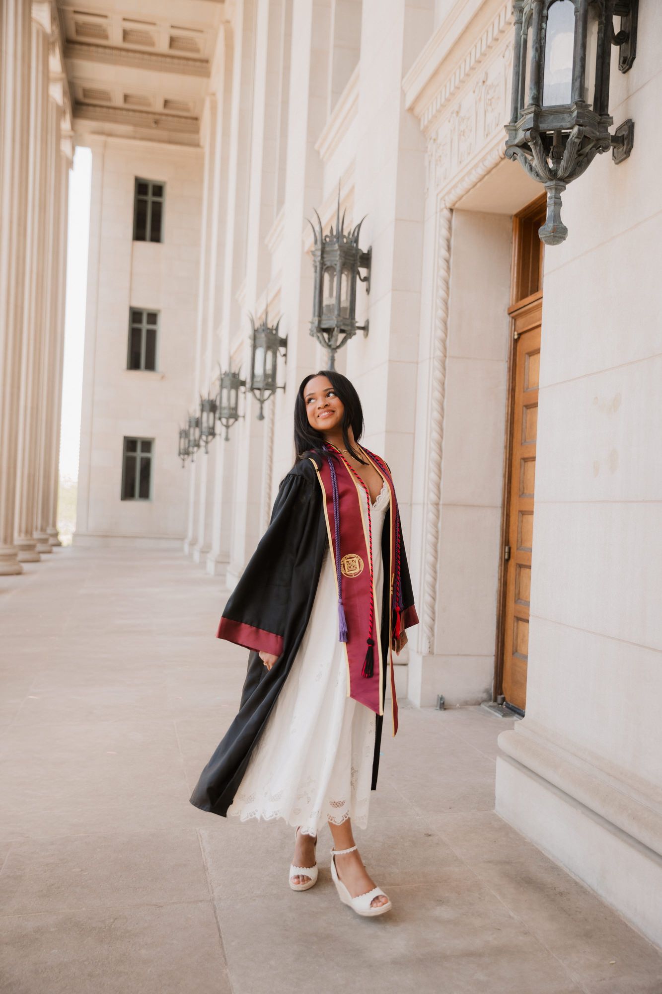 Young woman in black graduation gown and maroon stole poses between white columns of a university building.