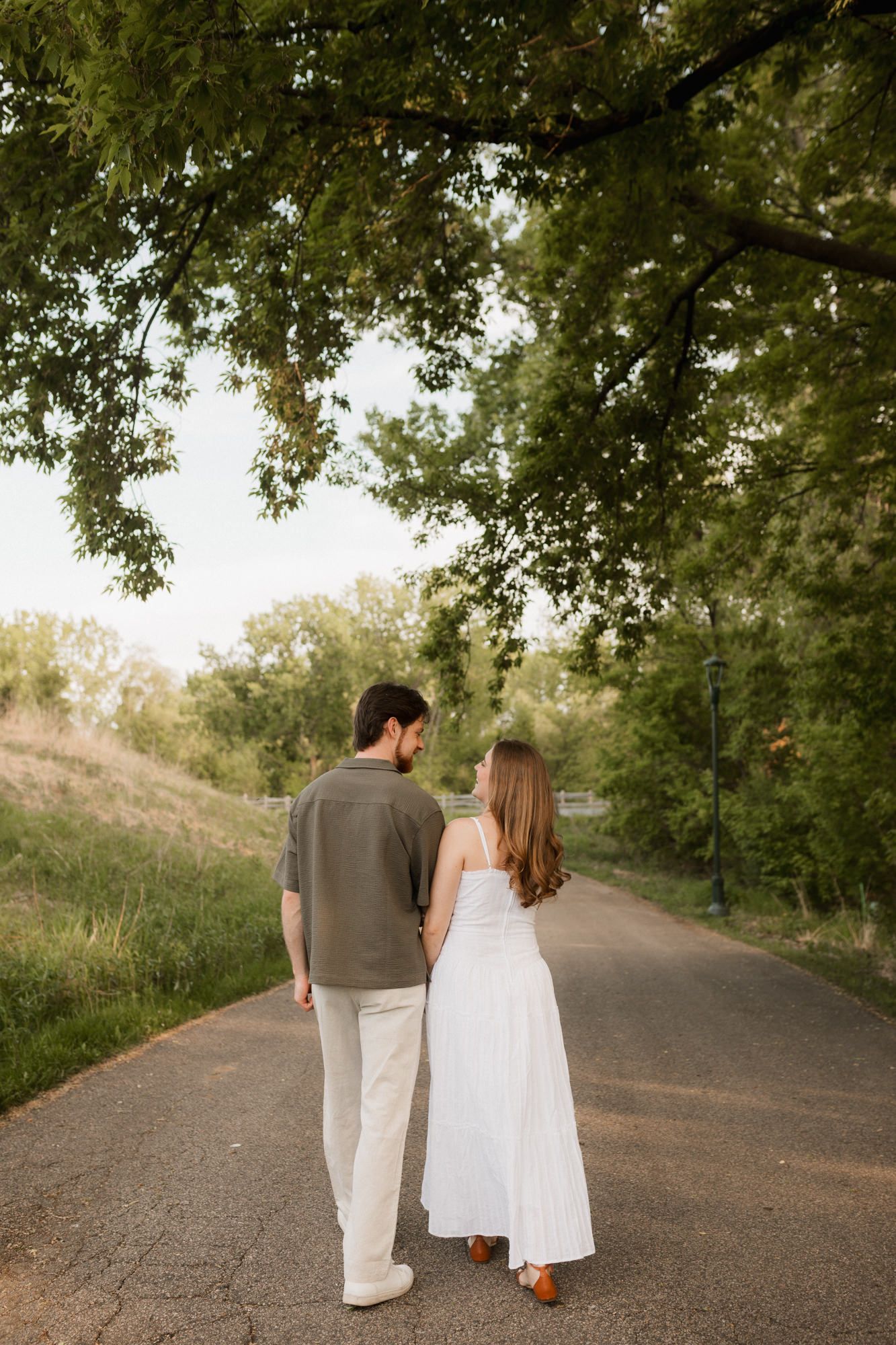Couple walking hand in hand on a tree-lined path, woman in white dress, man in olive shirt and white pants.