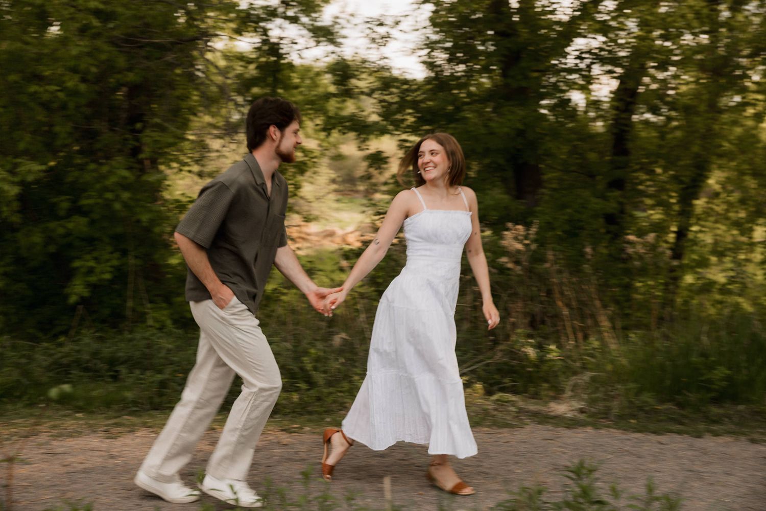 Couple holding hands and walking together outdoors in a lush green setting, woman in white dress, man in casual attire.