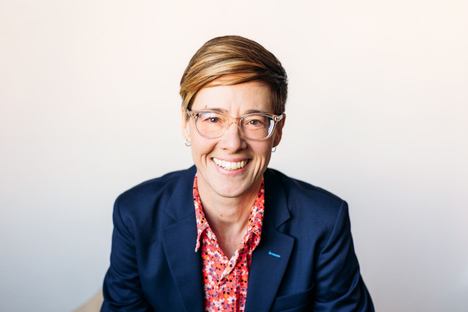 Smiling professional woman with short red hair and clear glasses wearing a navy blazer and floral shirt.