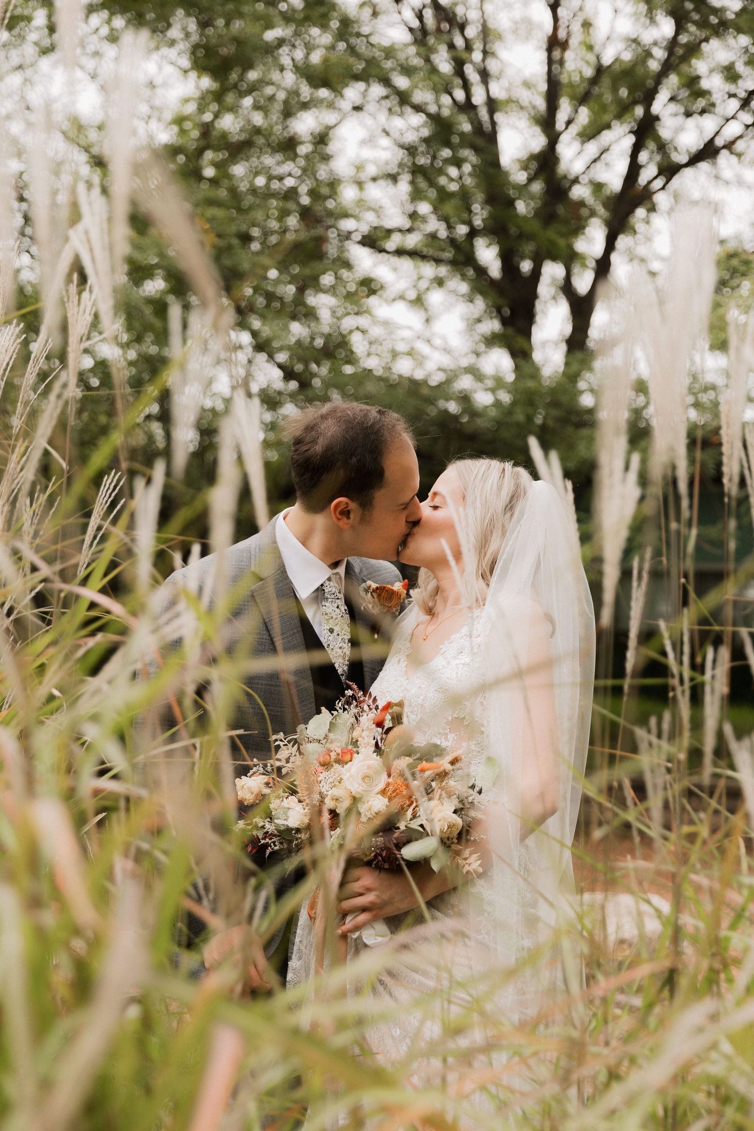 A bride and groom share a romantic kiss surrounded by tall ornamental grasses, bride holding a rustic bouquet.