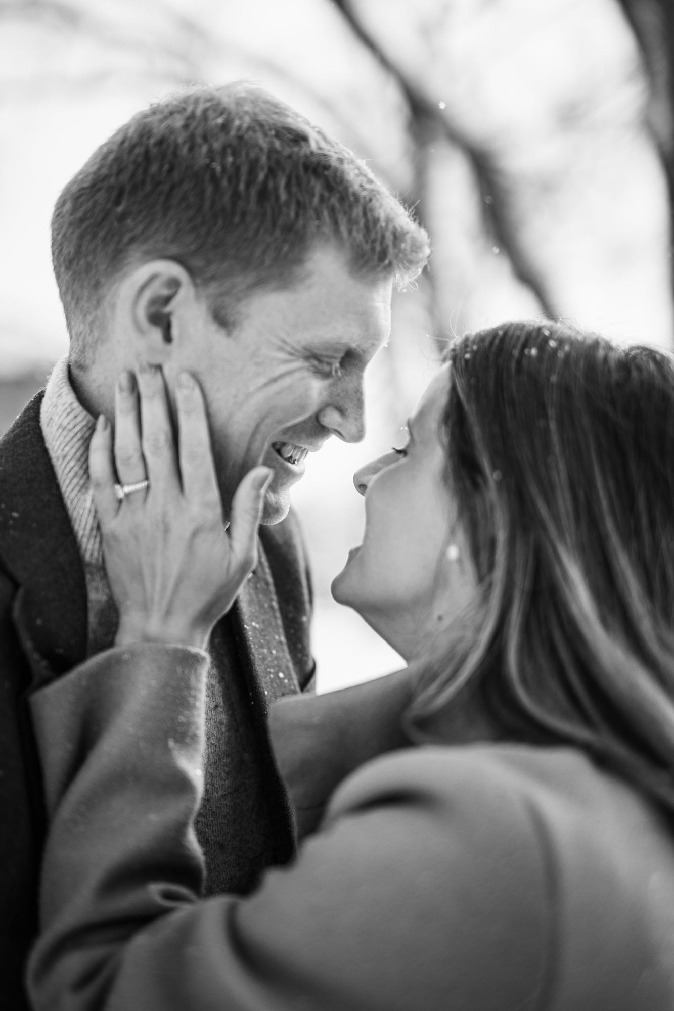Black and white photo of joyful couple touching foreheads, woman showing engagement ring in snow.