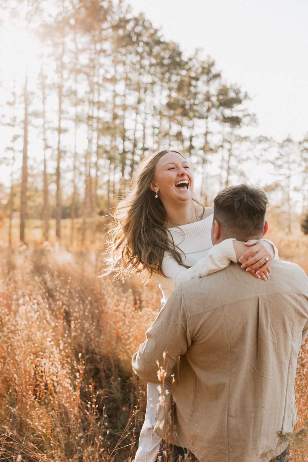 Joyful couple embraces in golden sunlit field with tall pine trees in background during sunset.