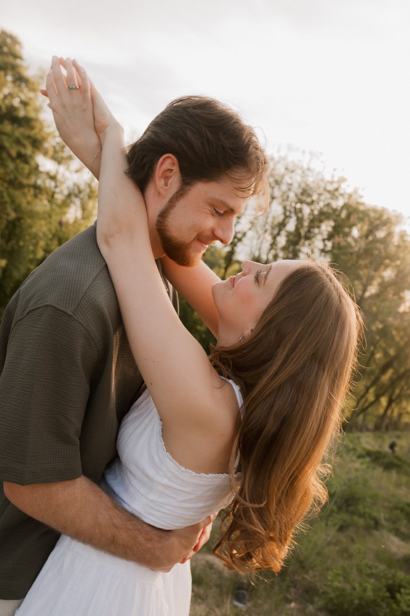 Couple embracing outdoors at golden hour, woman in white dress reaching up to mans face surrounded by trees.